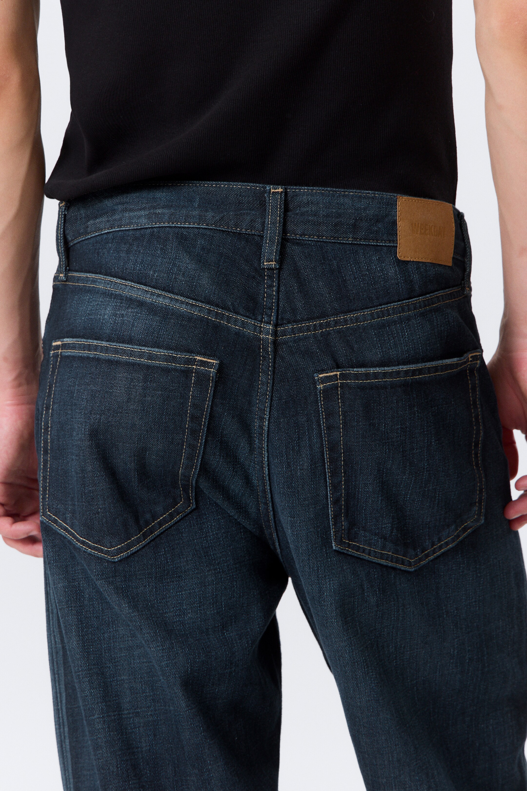 View larger image: Dark blue denim jeans are being presented from the back, showcasing their dark wash, prominent light brown contrast stitching on the two back patch pockets, and a brown leather-like brand patch visible on the center back waistband.