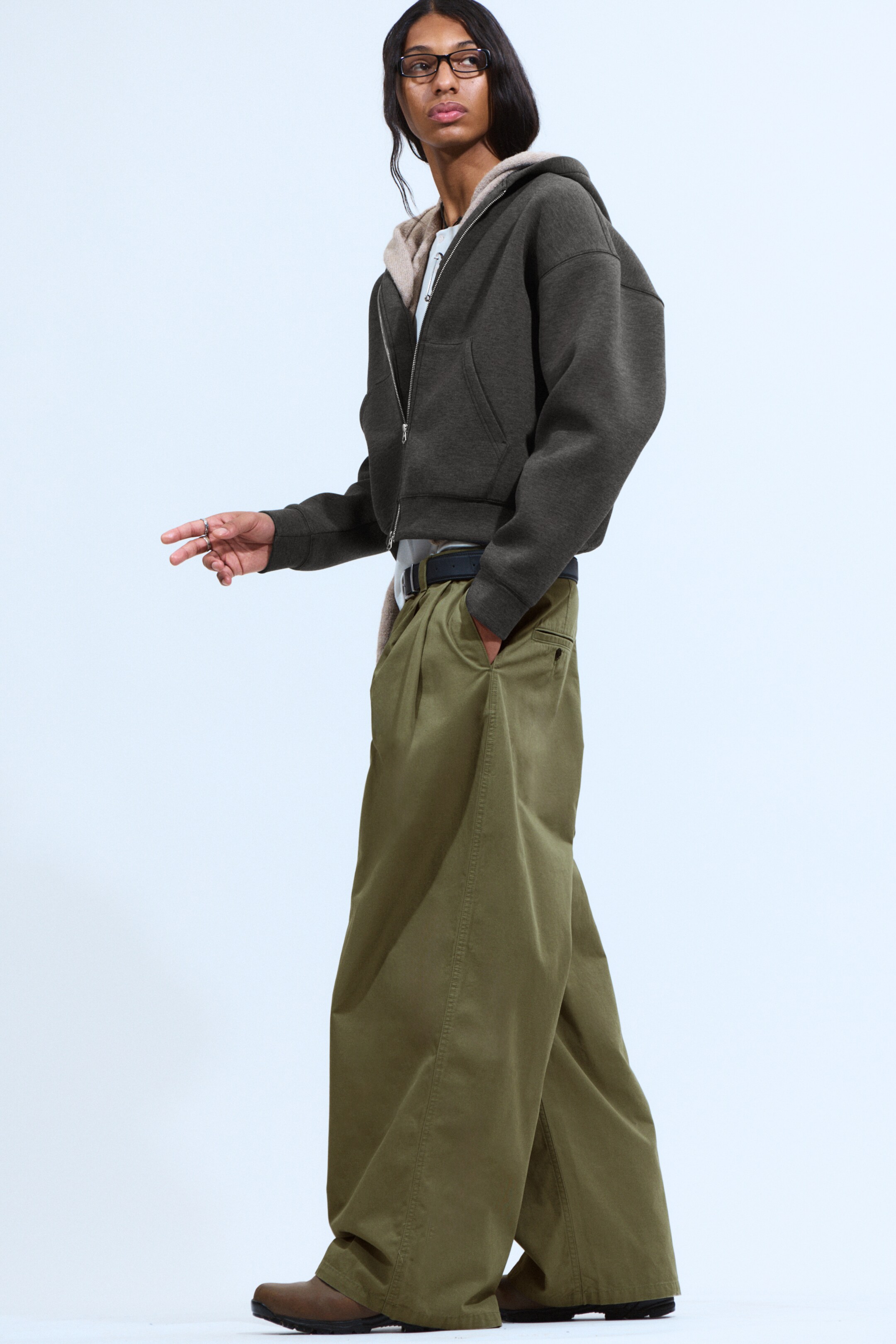 Agrandir l'image: Un pantalon chino vert olive surdimensionné, vu de la GAUCHE, présente une silhouette ample avec des jambes larges et une texture visible en sergé de coton. Ils sont conçus avec des plis avant proéminents, des poches latérales en biais et des passants de ceinture à travers lesquels une ceinture foncée est enfilée, ainsi qu'une fermeture à bouton supérieur.