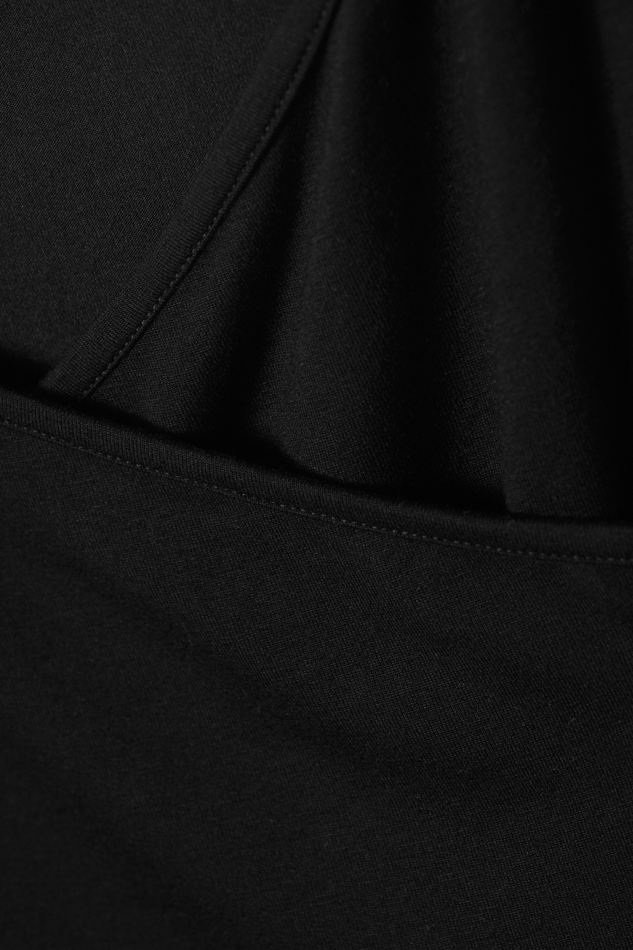 Größeres Bild anzeigen: Ein schwarzes Kleidungsstück zeigt eine Nahaufnahme seines Stoffes, mit einer diagonalen Naht mit sichtbaren Stichen und einer horizontalen genähten Kante, die ein überlappendes Detail erzeugt.