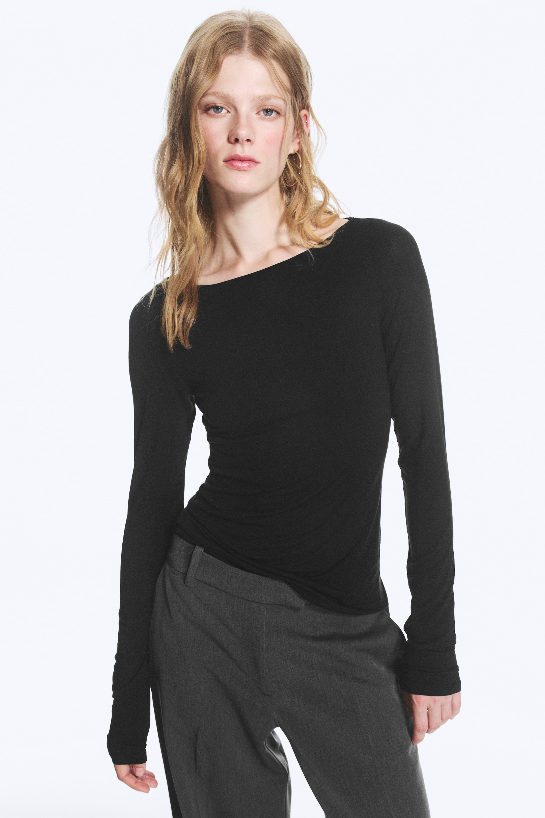 Visualizza l'immagine ingrandita: La modella indossa una maglia nera trasparente a maniche lunghe con scollo a barca. È aderente al busto, con il tessuto che si raccoglie intorno alla vita, abbinata a pantaloni grigi.