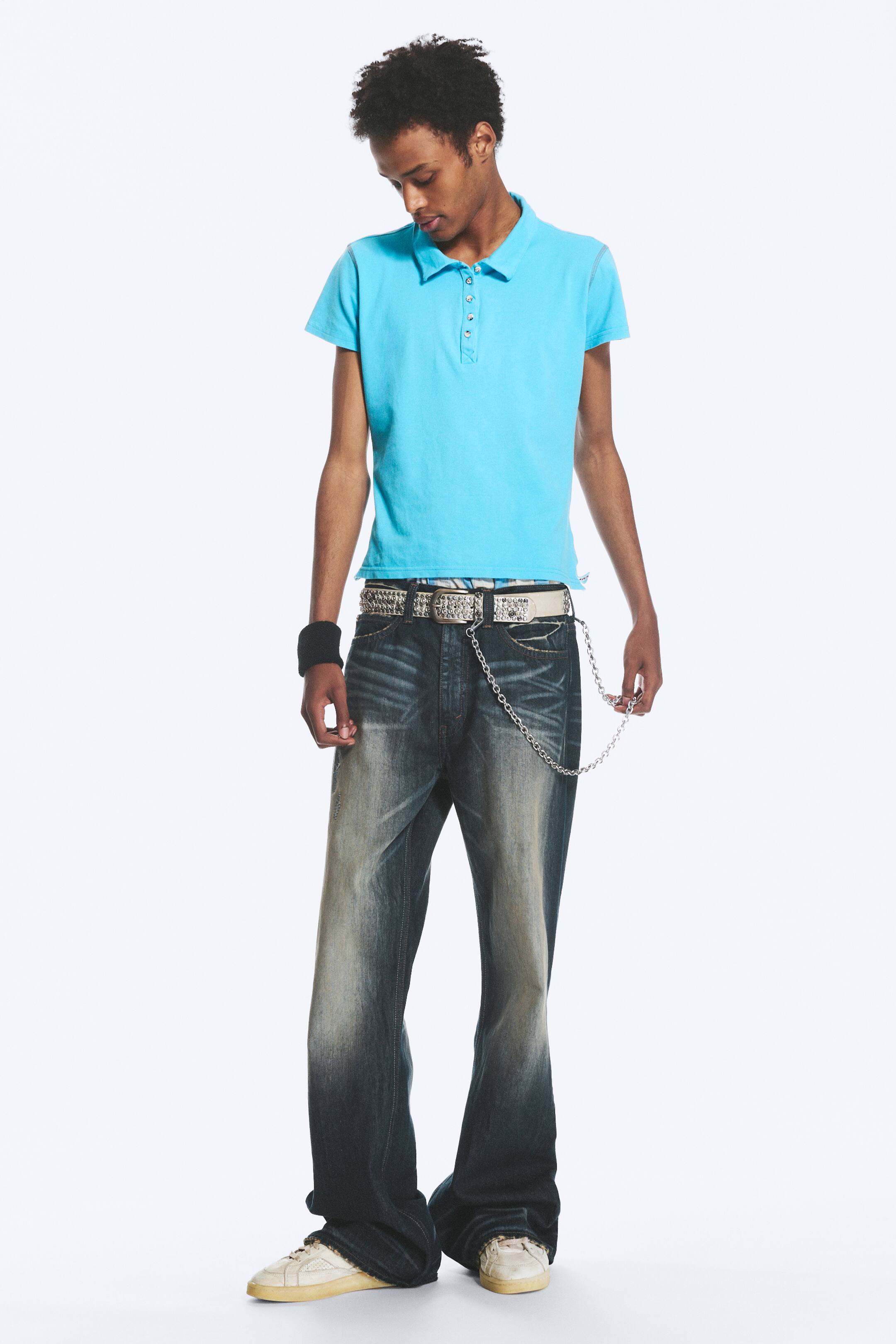 Zobacz większy obraz: Model ma na sobie jeansy z niskim stanem i fasonem bootcut w spranym, ciemnym odcieniu, zestawione z jasnoniebieską koszulką polo. Jeansy wystylizowano z paskiem z ćwiekami oraz srebrnym metalowym łańcuchem zwisającym z szlufek, opadającym na jasne sneakersy.