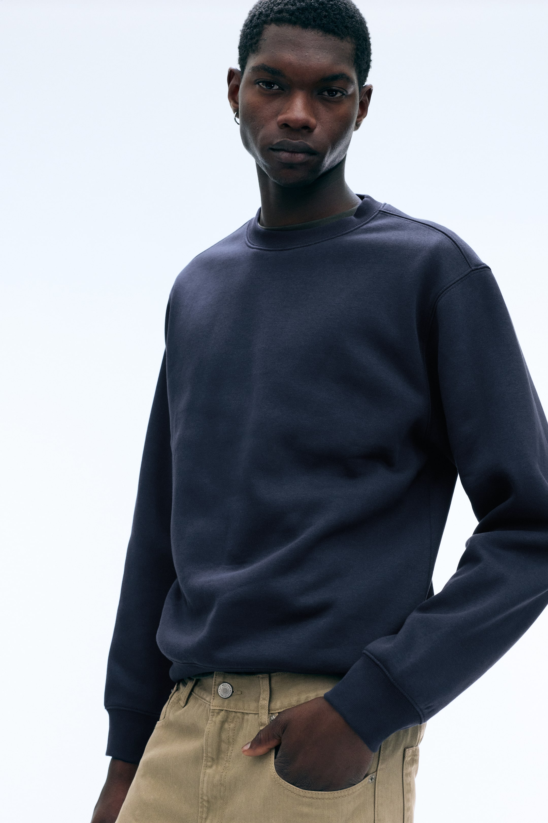 Visa större bild: En mörk marinblå crewneck-tröja med nedhasade axlar och ribbade muddar bärs av modellen. Tröjan är stylad med beige byxor, och modellen vilar ena handen i en framficka.