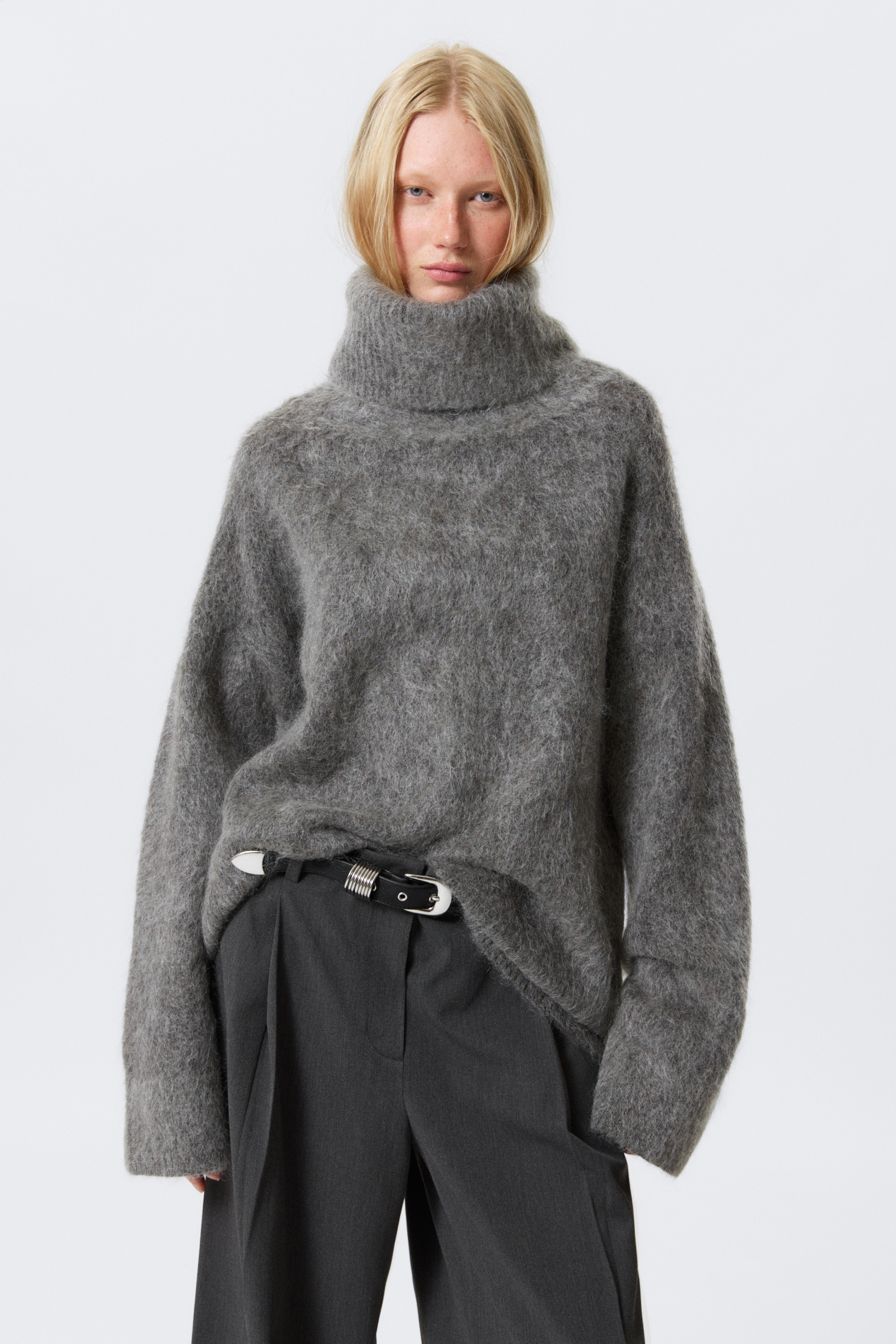 Grotere afbeelding bekijken: Een grijze, pluizige oversized coltrui wordt van voren gedragen. Het heeft een hoge, losse colkraag en een ontspannen pasvorm met lange mouwen, waarbij de zoom nonchalant in een donkere broek is gestopt, waardoor een zwarte riem met zilverkleurige details zichtbaar wordt.