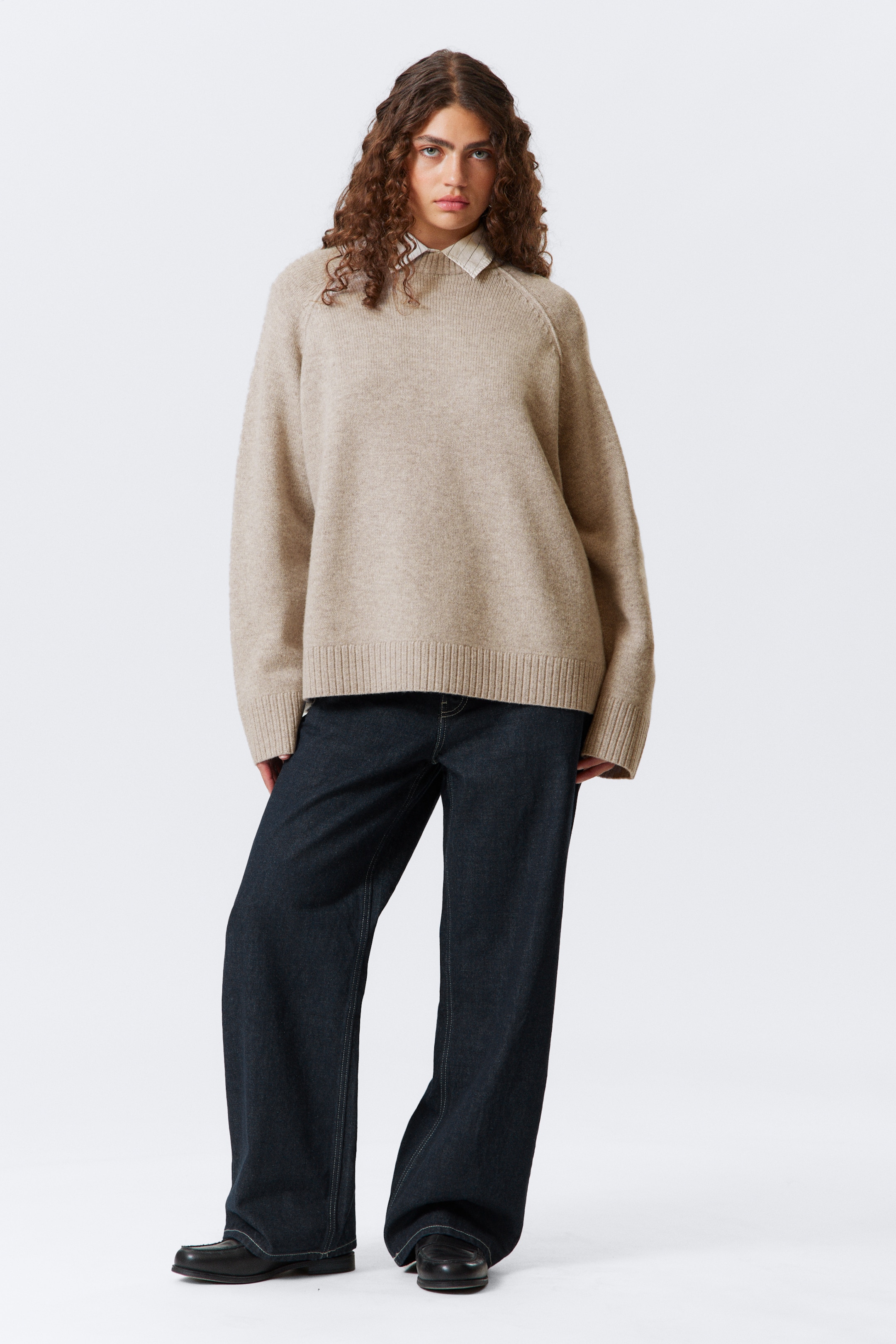 Größeres Bild ansehen: Ein beige gestrickter Pullover mit Rundhalsausschnitt wird getragen, der eine entspannte, übergroße Passform, Raglanärmel und gerippte Bündchen und einen gerippten Saum aufweist. Ein weißes Hemd mit Kragen ist sichtbar unter dem Ausschnitt des Pullovers geschichtet.