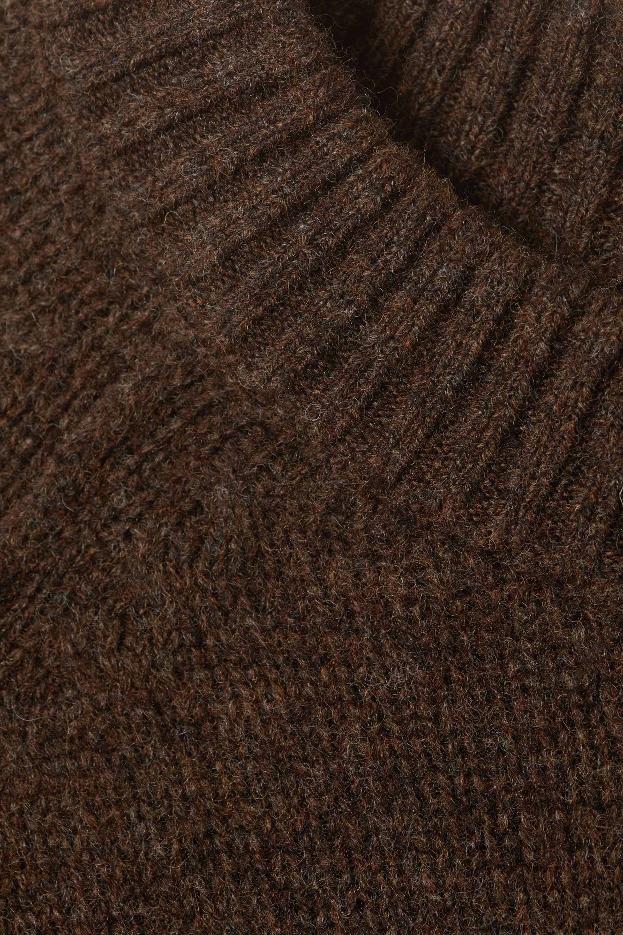 Agrandir l'image: Un tissu tricoté brun foncé présente une surface texturée avec des fibres visibles, où une section côtelée, possiblement un col ou un poignet, se replie sur le corps principal du tricot, formant une forme en V et montrant des côtes verticales distinctes.