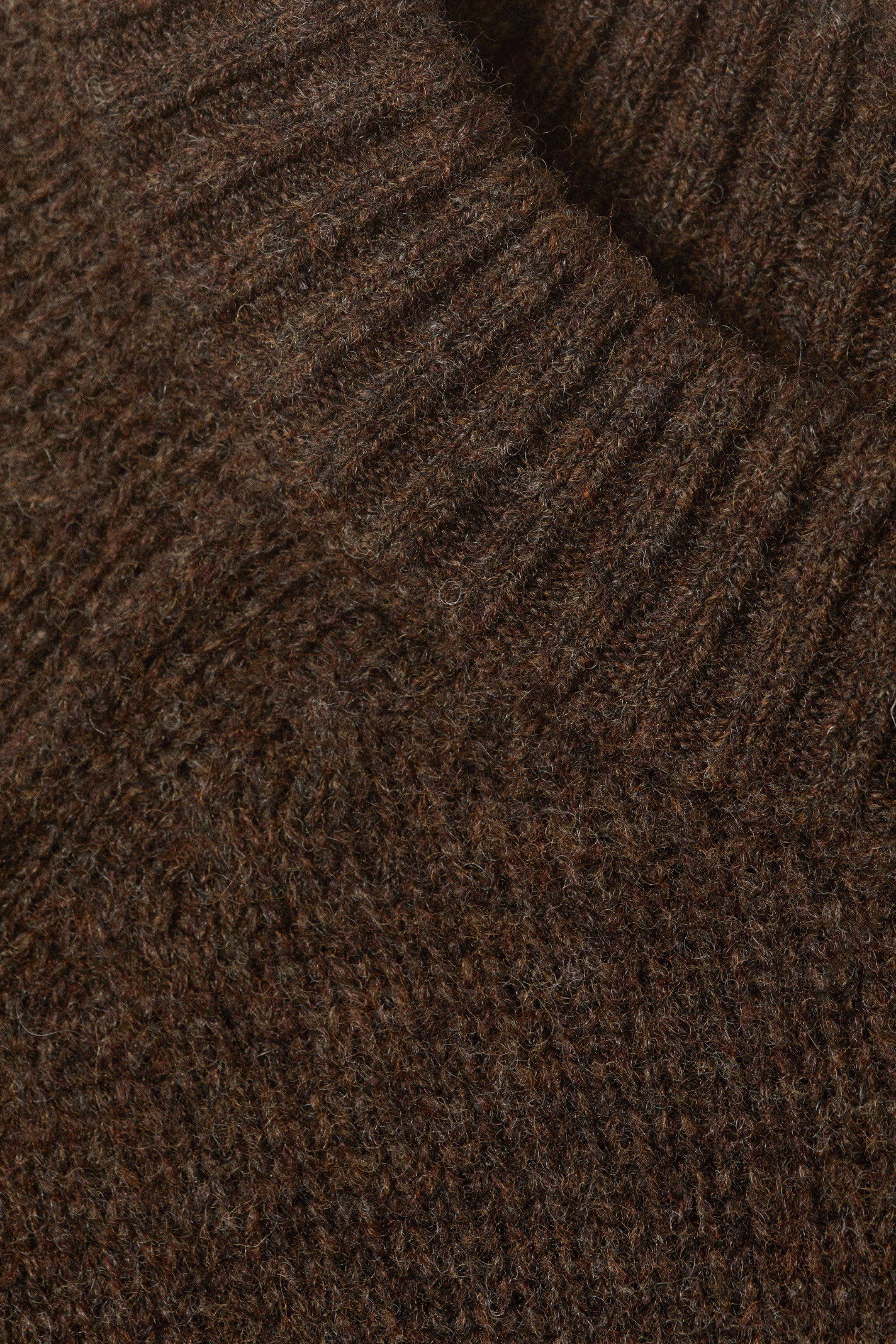 Größeres Bild anzeigen: Ein dunkelbrauner Strickstoff präsentiert eine strukturierte Oberfläche mit sichtbaren Fasern, wobei ein gerippter Abschnitt, möglicherweise ein Kragen oder eine Manschette, über den Hauptteil des Stricks umgeschlagen ist und eine V-Form bildet, die deutliche vertikale Rippen zeigt.