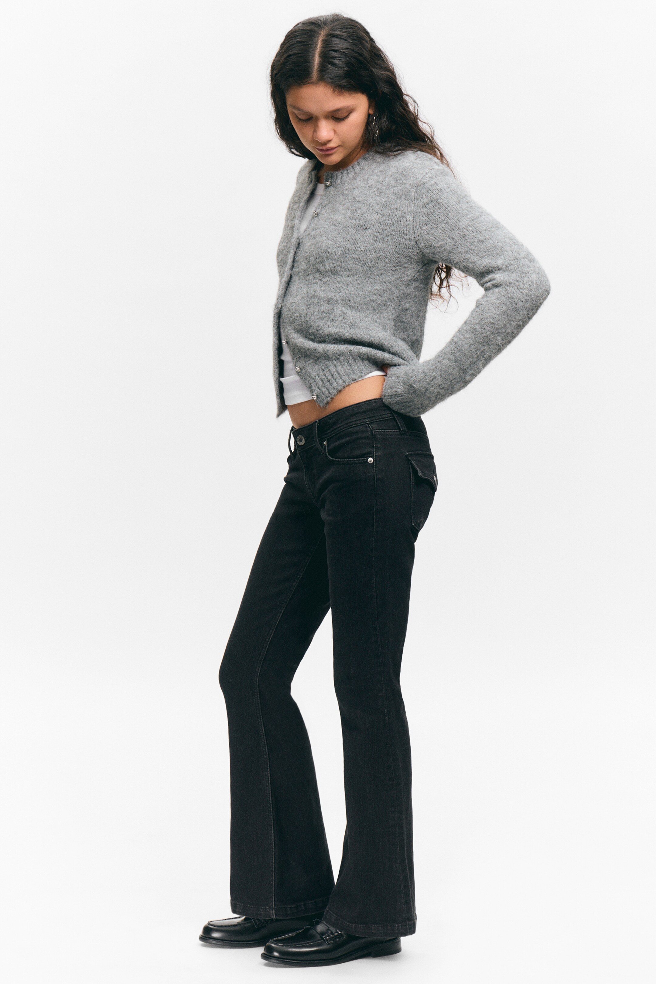 Agrandir l'image: Un cardigan court en tricot gris duveteux, avec des poignets et un ourlet côtelés et des boutons ronds à l'avant, est porté ouvert sur un haut blanc et associé à un jean évasé noir et des mocassins noirs, le tout présenté sous un angle de vue de côté droit.
