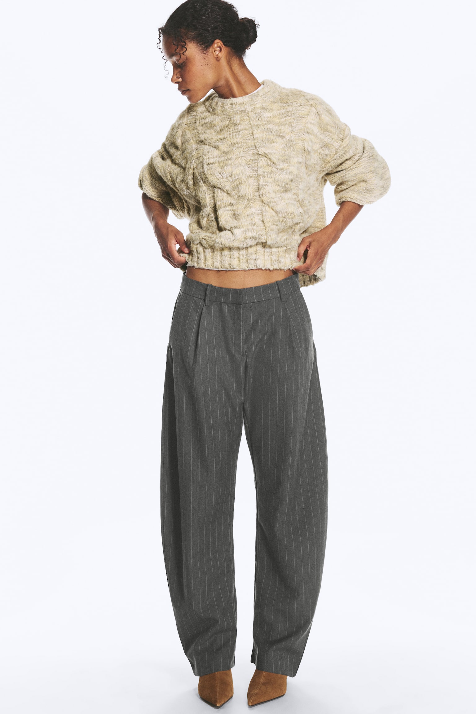 Das Model trägt eine graue Anzughose mit Nadelstreifen, lockerem Schnitt und barrel-förmigem Bein. Es hebt den Saum eines beigefarbenen Zopfstrickpullovers an, um den mittelhohen Bund der Hose sichtbar zu machen. - 1