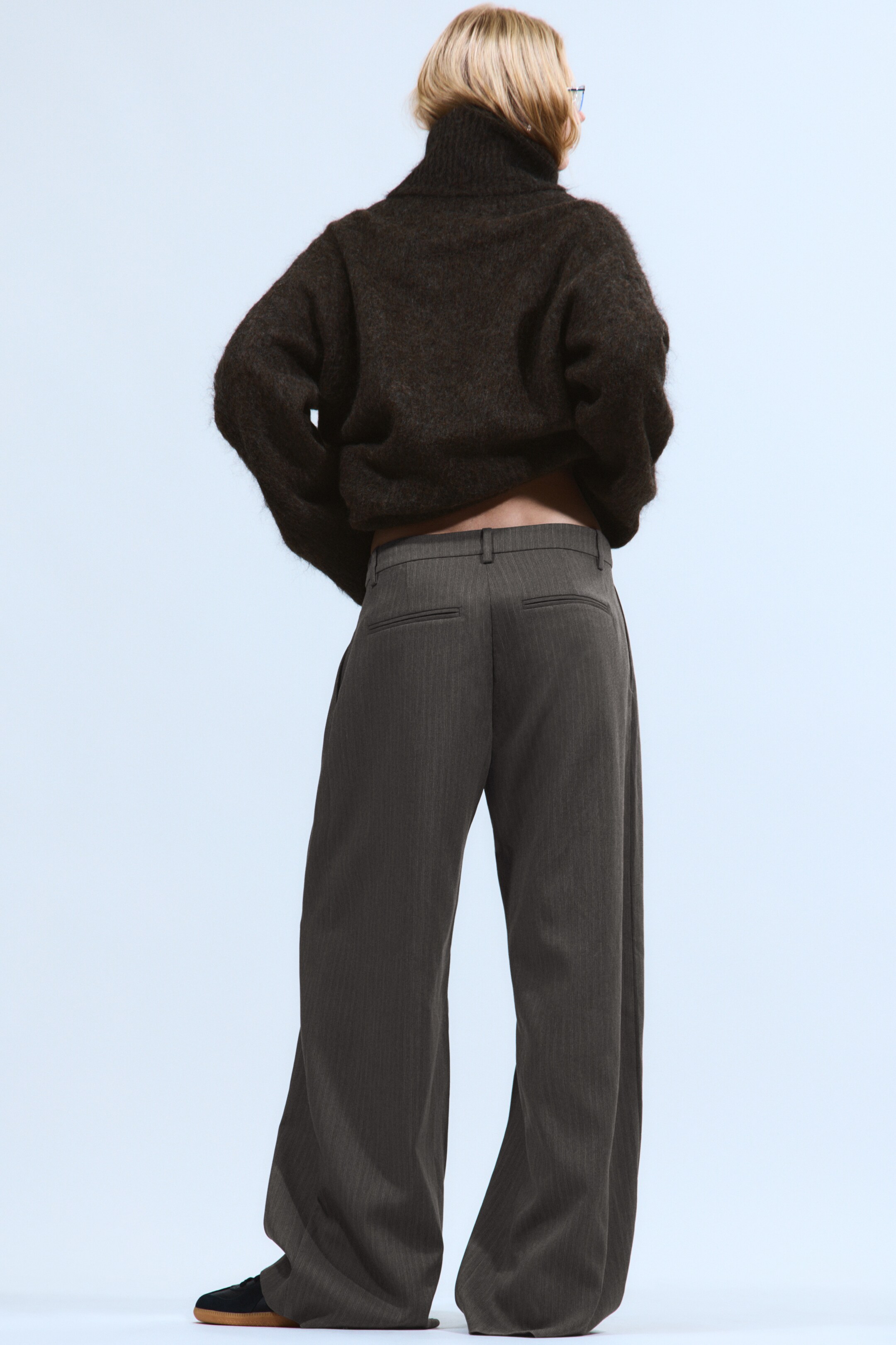 Ingrandisci l'immagine: Un maglione a collo alto a maglia grossa di colore marrone scuro è mostrato dal retro, abbinato a pantaloni a gamba larga con righe sottili di colore grigio scuro. Il maglione mostra una vestibilità ampia e corta con un collo alto, mentre i pantaloni presentano una silhouette a gamba larga a tutta lunghezza che si raccoglie sul fondo e mostrano due tasche posteriori visibili.