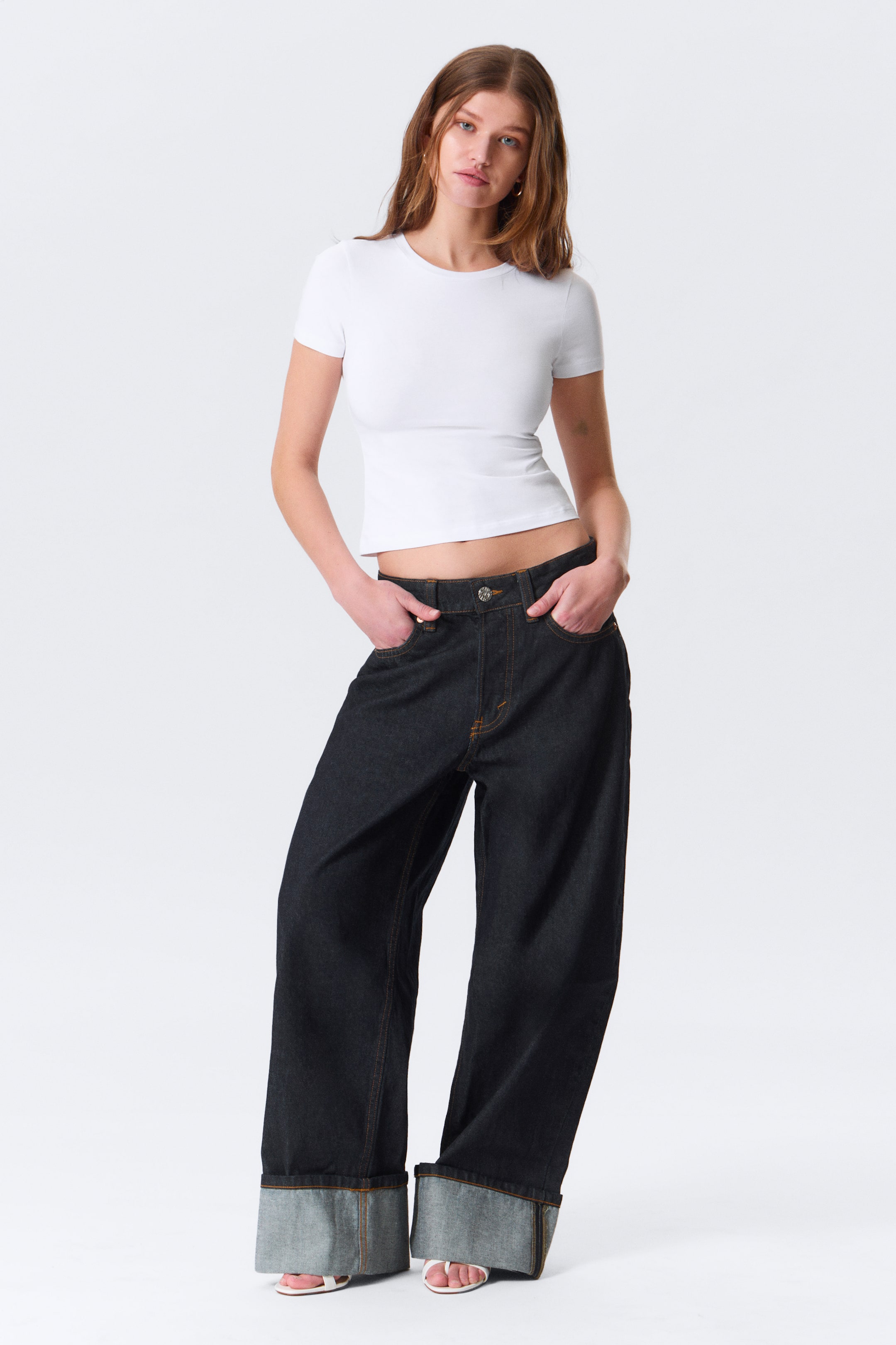 Grösseres Bild ansehen: Ein weißes, eng anliegendes T-Shirt mit kurzen Ärmeln und Rundhalsausschnitt wird mit dunkel gewaschenen, weit geschnittenen Jeans kombiniert, die durch kontrastierende Nähte und auffällige umgeschlagene Säume mit einem helleren Denim-Innenfutter auffallen.