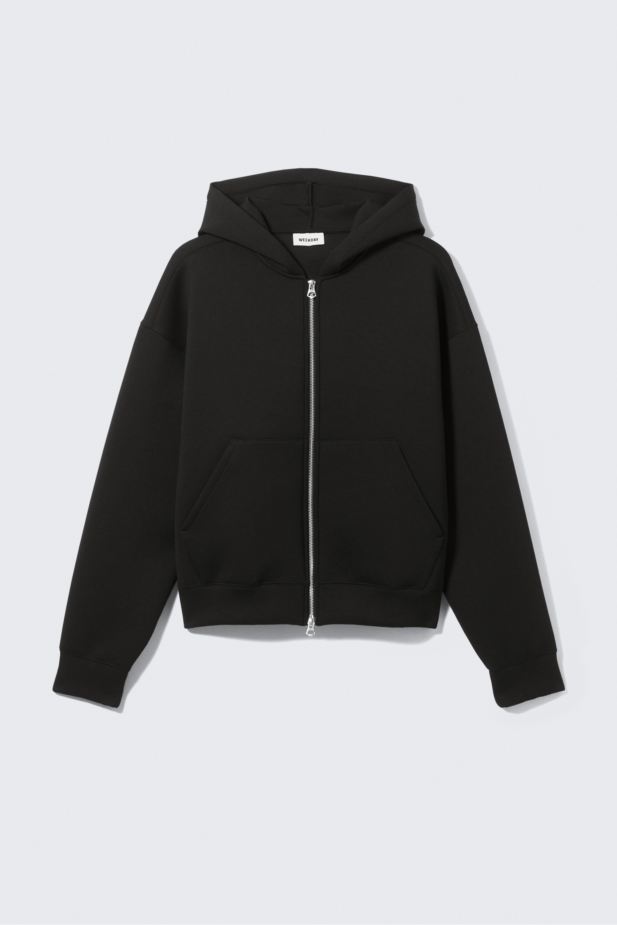 View larger image: A black zip-up hoodie is being presented, facing forward. It is featuring a silver-toned central zipper, two large angled patch pockets, and a relaxed fit with drop shoulders, ribbed cuffs, and a ribbed hem. A small white 'WEEKDAY' brand tag is visible inside the hood.