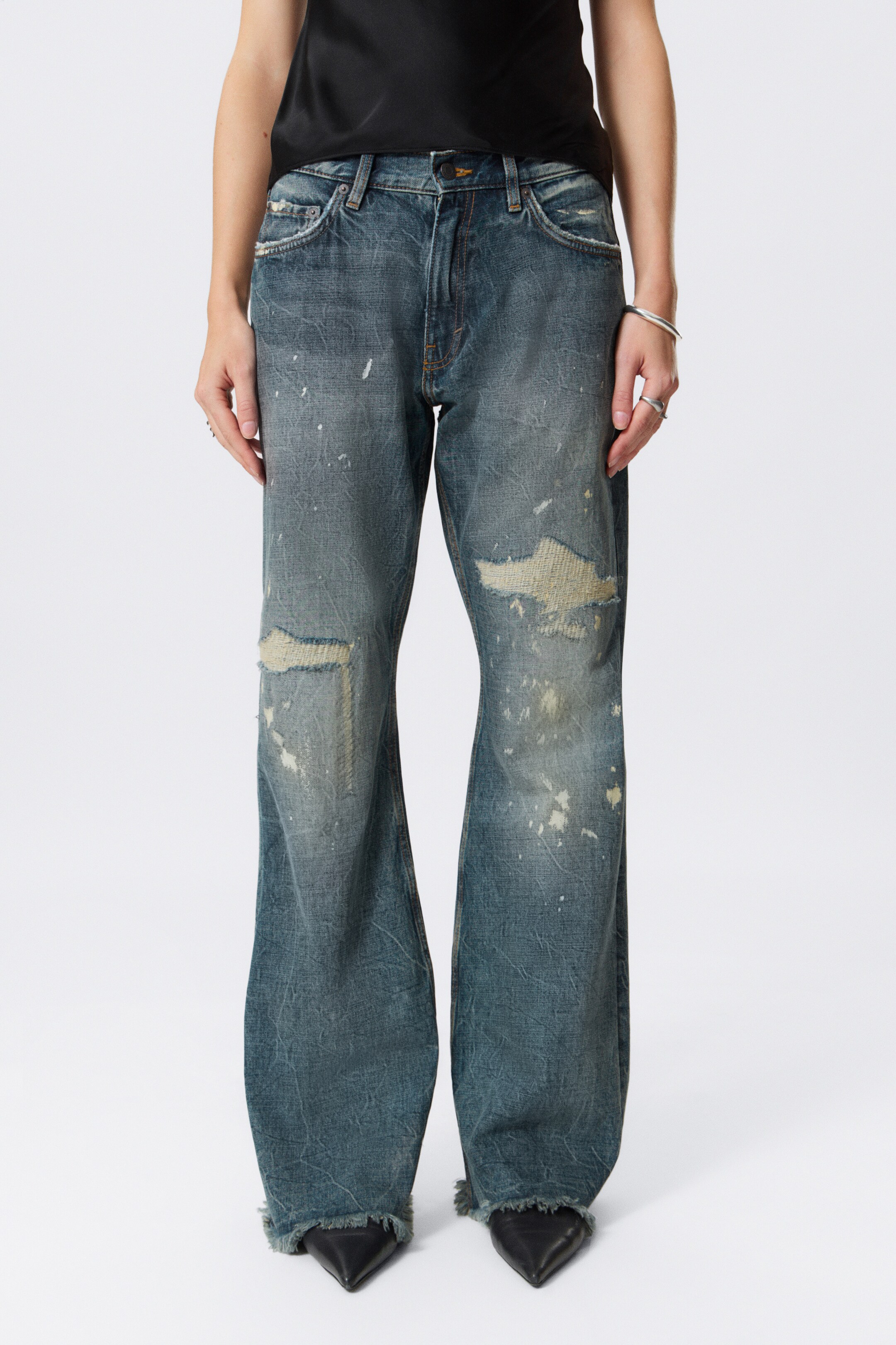 Größeres Bild anzeigen: Ein Paar ausgewaschene blaue Denim-Jeans mit niedriger Taille, ausgestelltem Bein und rohen Säumen wird von dem Modell getragen. Die Jeans weist starke Abrasionen an der Vorderseite und Farbspritzer auf und wird mit schwarzen spitzen Schuhen gestylt.