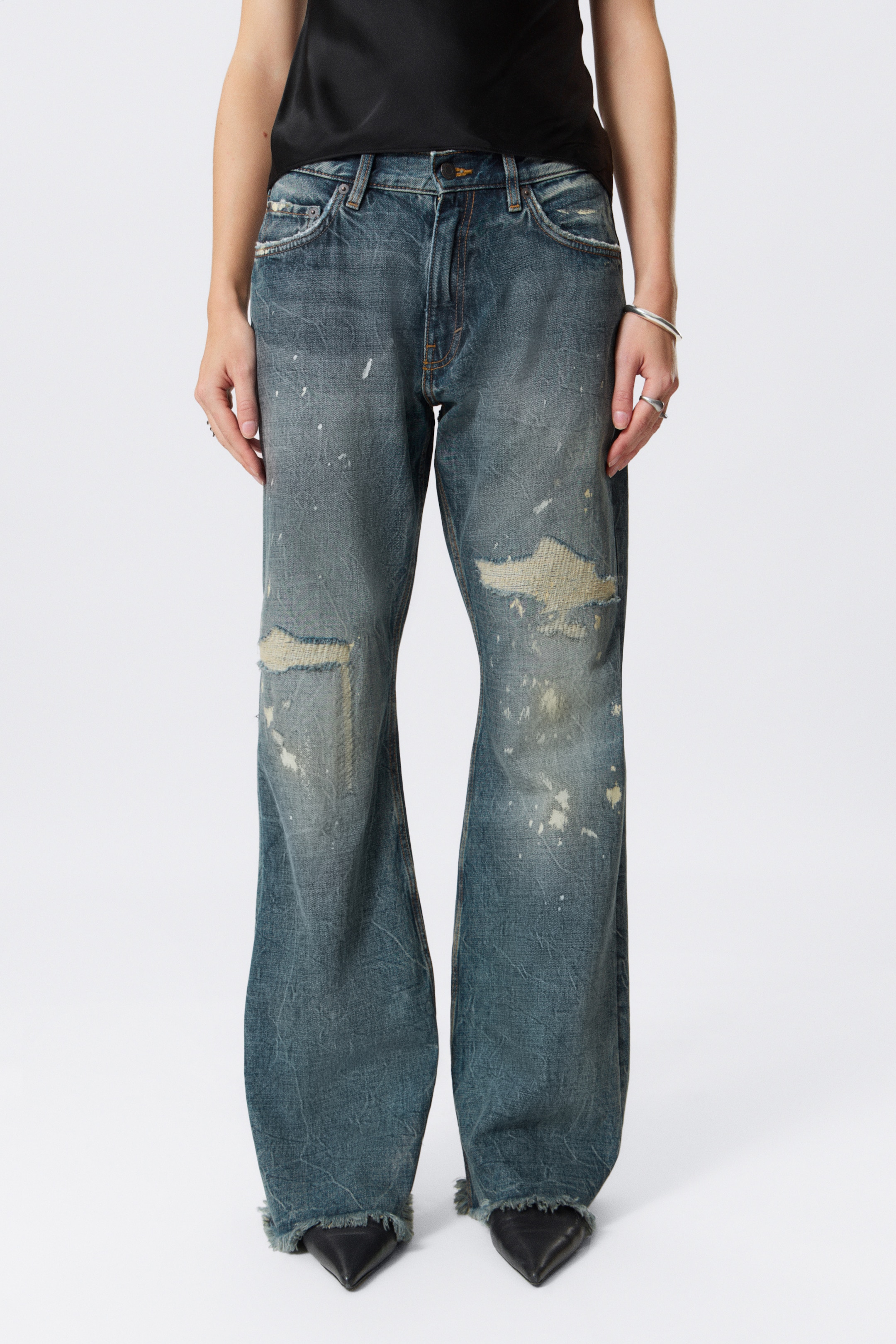 Agrandir l'image: Un jean en denim bleu délavé avec une taille basse, une jambe évasée, et des ourlets bruts est porté par le mannequin. Le jean présente de fortes abrasions sur le devant et des éclaboussures de peinture, et est assorti de chaussures noires à bout pointu.