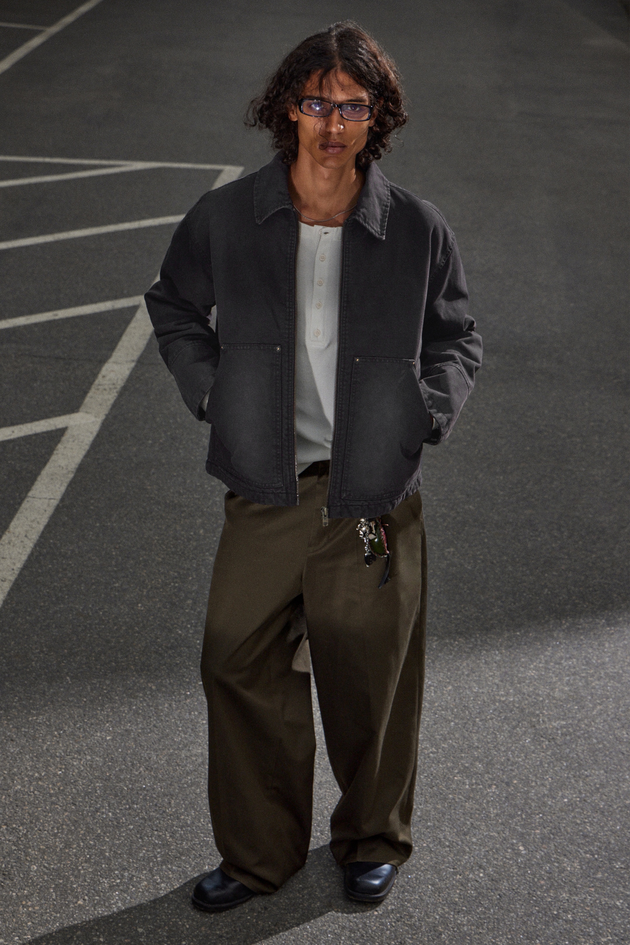 View larger image: A dark gray workwear jacket is being worn over a light-colored henley shirt, paired with olive wide-leg pants and black shoes. The jacket is featuring a classic collar and two large patch pockets with visible rivets on the lower front, while keychains are hanging from the front of the pants.