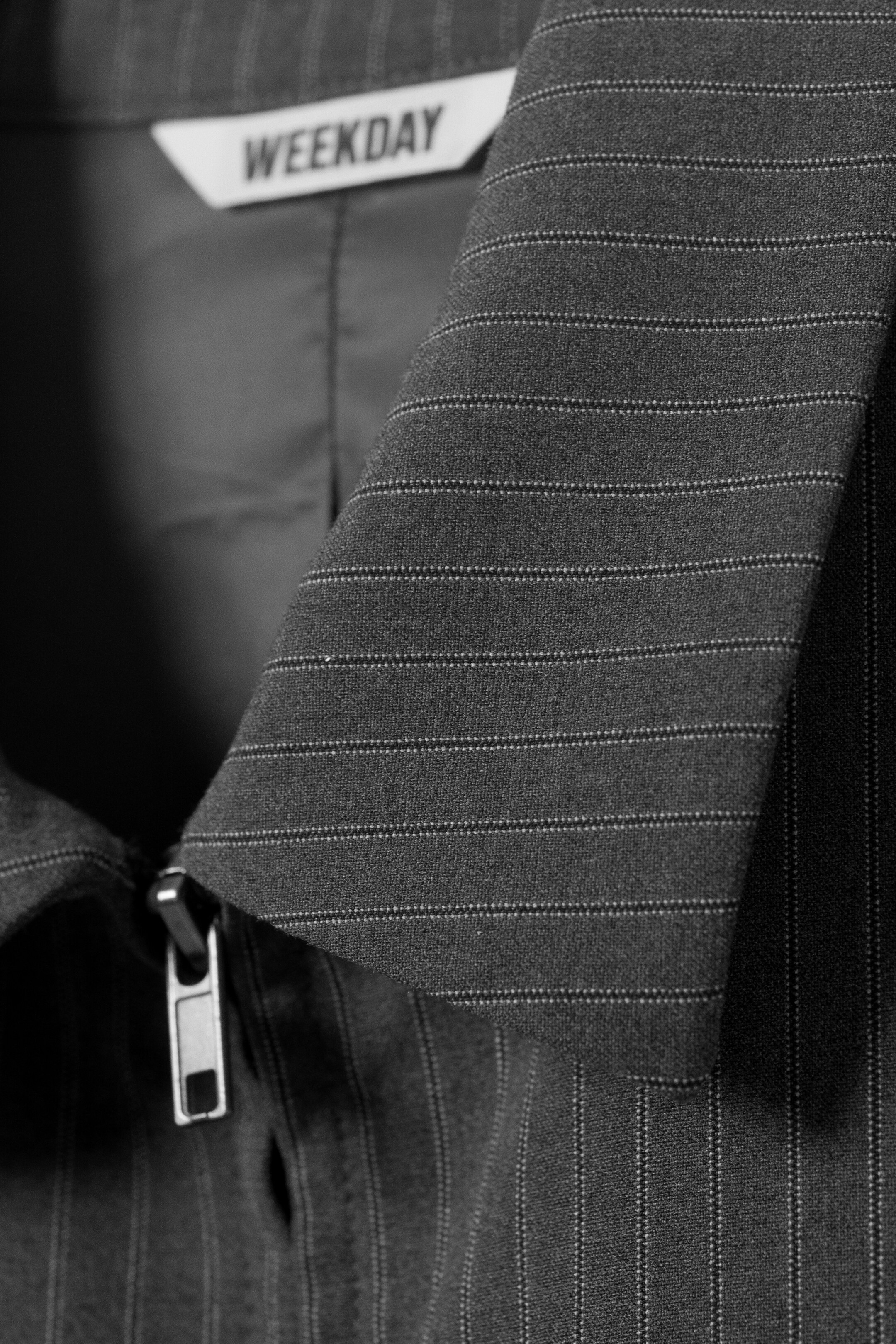 Agrandir l'image: Une veste à rayures fines gris foncé présente un col structuré et une tirette de fermeture éclair argentée sur le devant supérieur. Une étiquette blanche "WEEKDAY" est visible à l'intérieur du cou.
