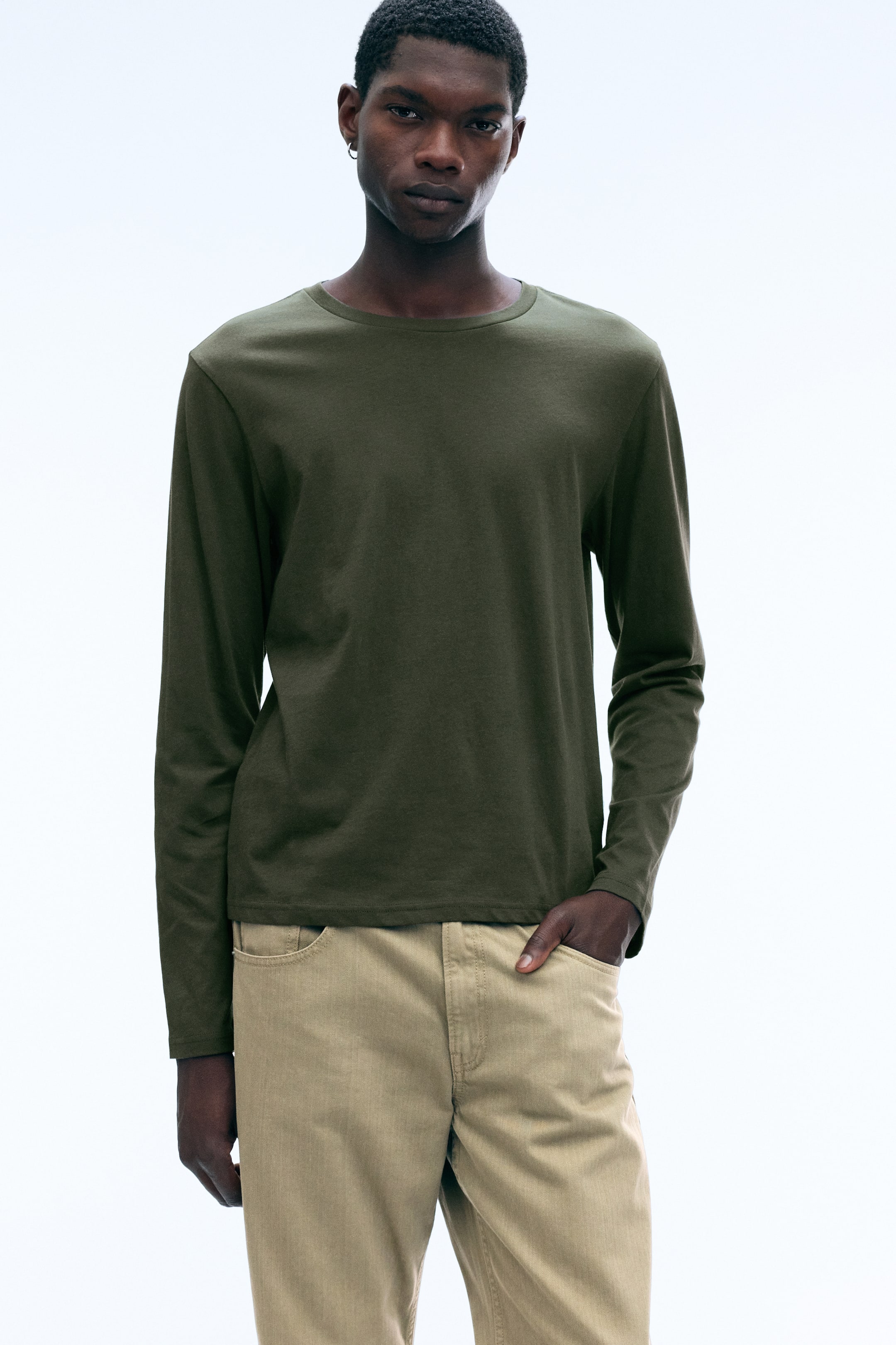 Agrandir l'image: Un T-shirt à manches longues vert olive et à col rond est porté par le mannequin, associé à un pantalon beige. Le mannequin est debout, une main glissée dans une poche avant.