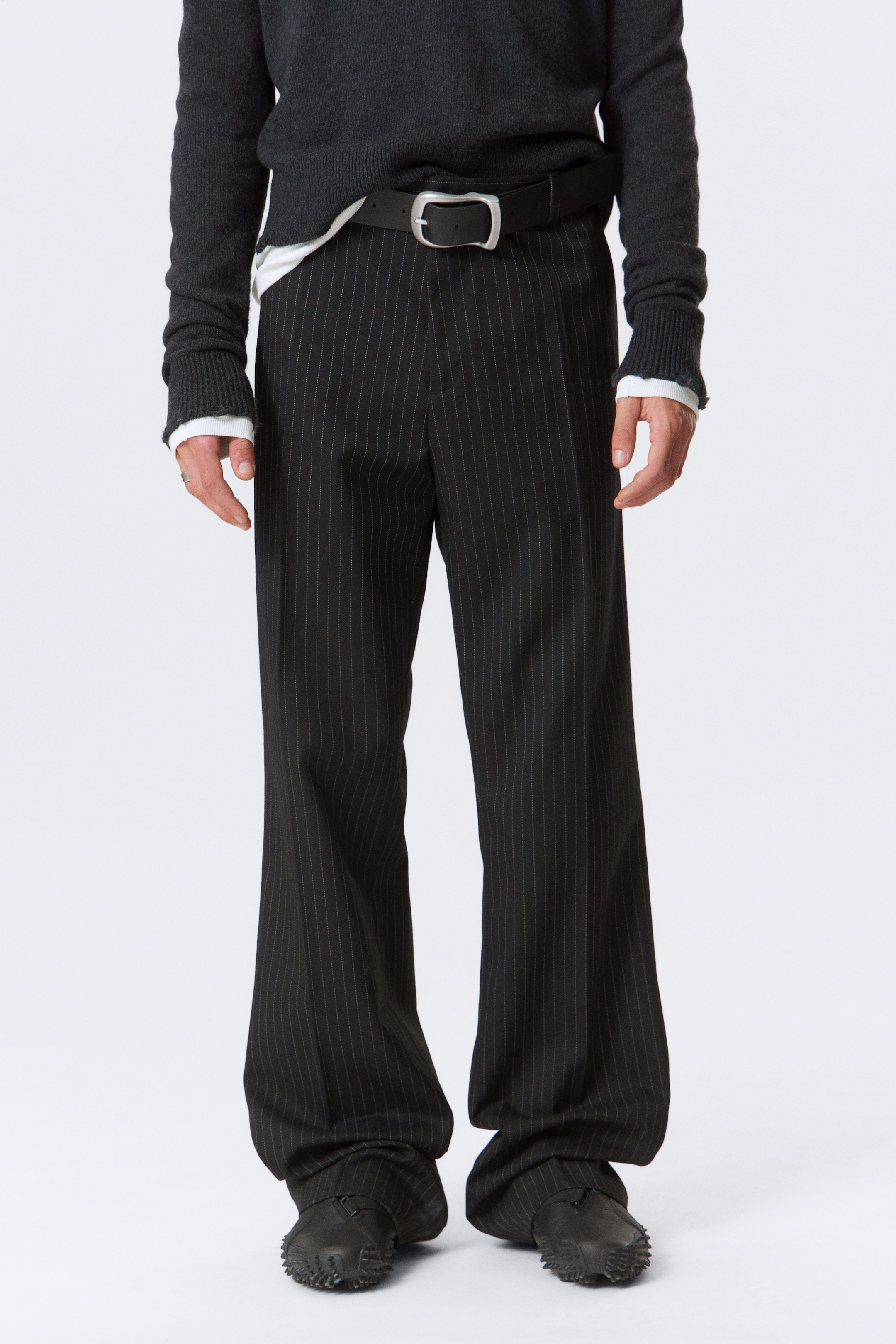Agrandir l'image: Le mannequin porte un pantalon noir à rayures, présentant une coupe large. Une ceinture noire avec une boucle argentée sécurise le pantalon à la taille, et les ourlets se regroupent sur des chaussures noires texturées.