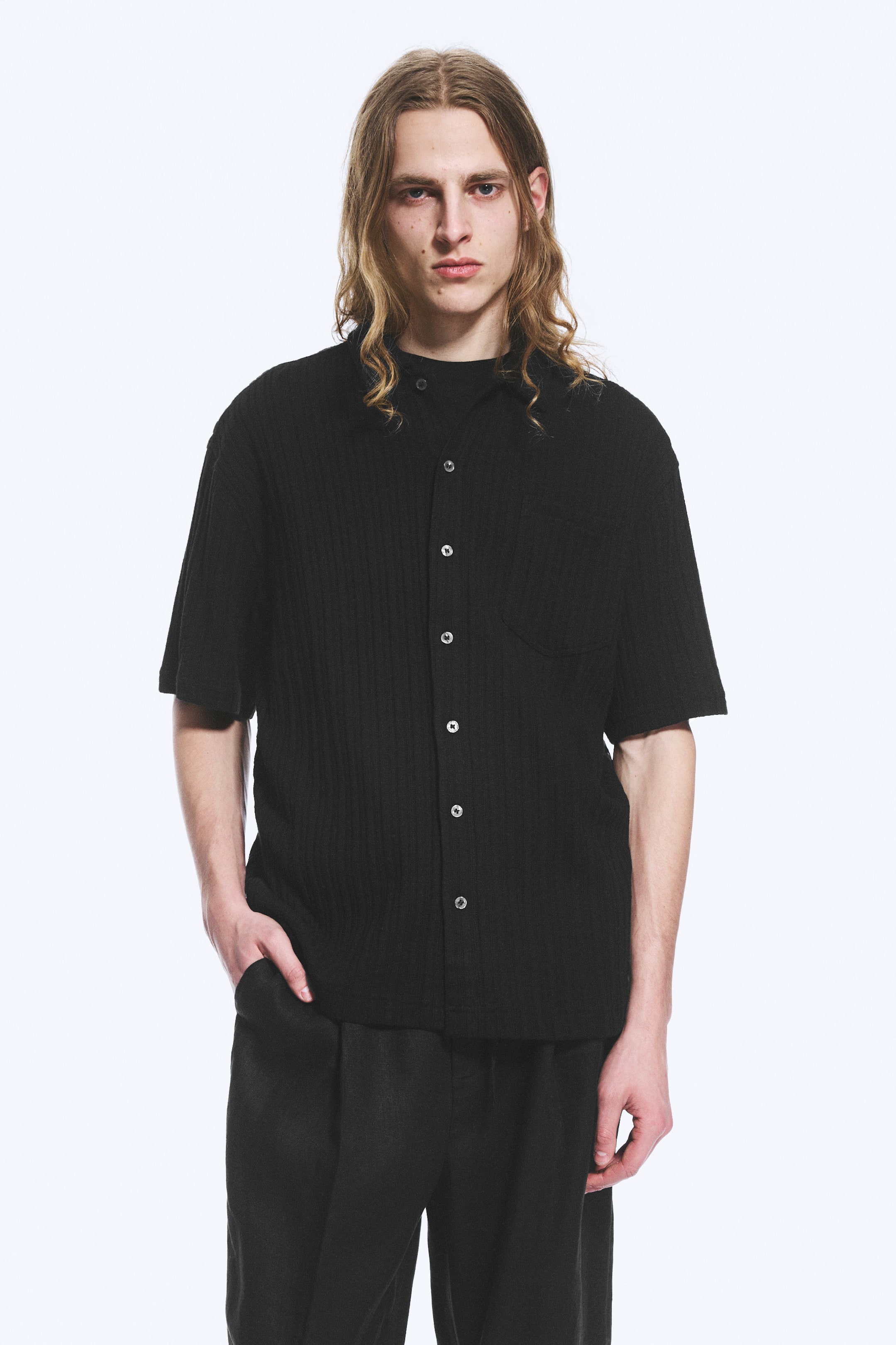 Ver imagen más grande: Una camisa negra de manga corta está orientada hacia adelante, con una textura acanalada, un cuello en punta y un cierre frontal de botones con botones de color claro. Se aprecia un bolsillo en el pecho en el lado izquierdo, y la camisa está superpuesta sobre una camiseta interior oscura.