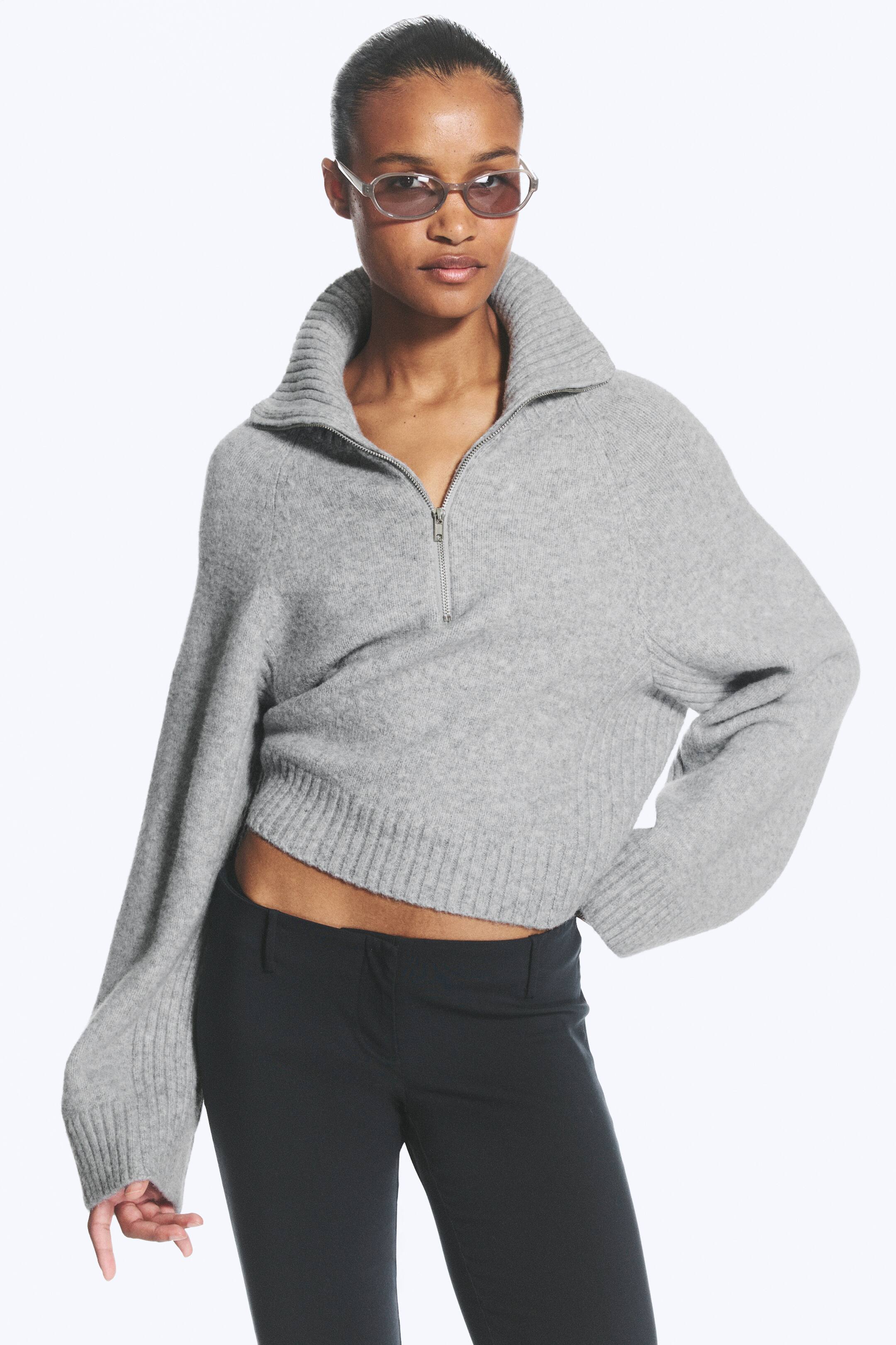 Größeres Bild ansehen: Der graue Cropped-Pullover mit Viertelreißverschluss verfügt über einen gerippten Stehkragen und einen silberfarbenen Reißverschluss, der derzeit teilweise geöffnet ist. Er zeigt lange Raglanärmel mit gerippten Bündchen sowie einen gerippten Saum, der oberhalb der Taille endet.