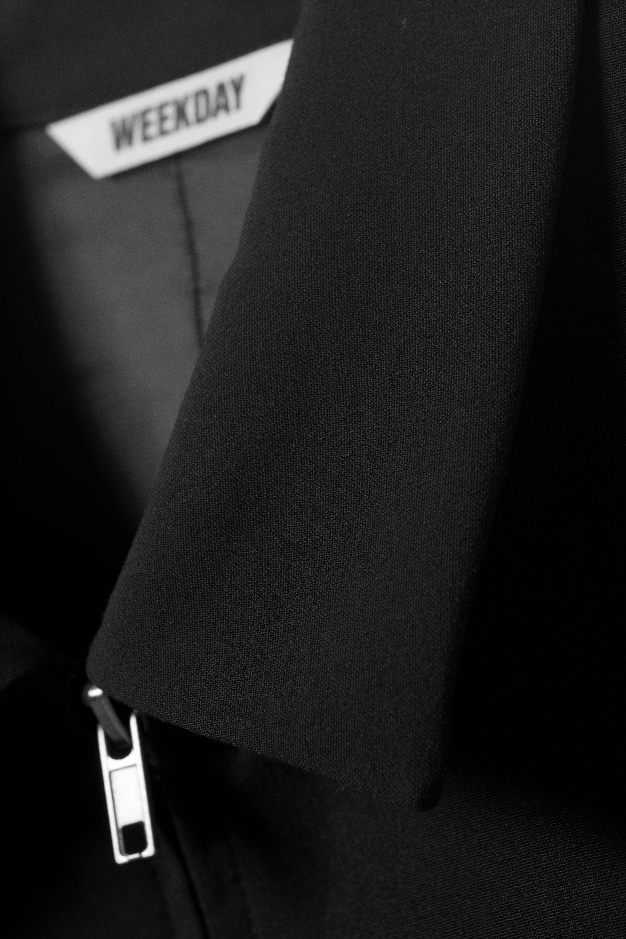 Größeres Bild anzeigen: Eine Nahaufnahme einer schwarzen Jacke zeigt ein strukturiertes Revers oder einen Kragen, mit einem silbernen Reißverschlussgriff, der unten links sichtbar ist, und einem weißen "WEEKDAY"-Markenetikett, das innen oben links eingenäht ist.