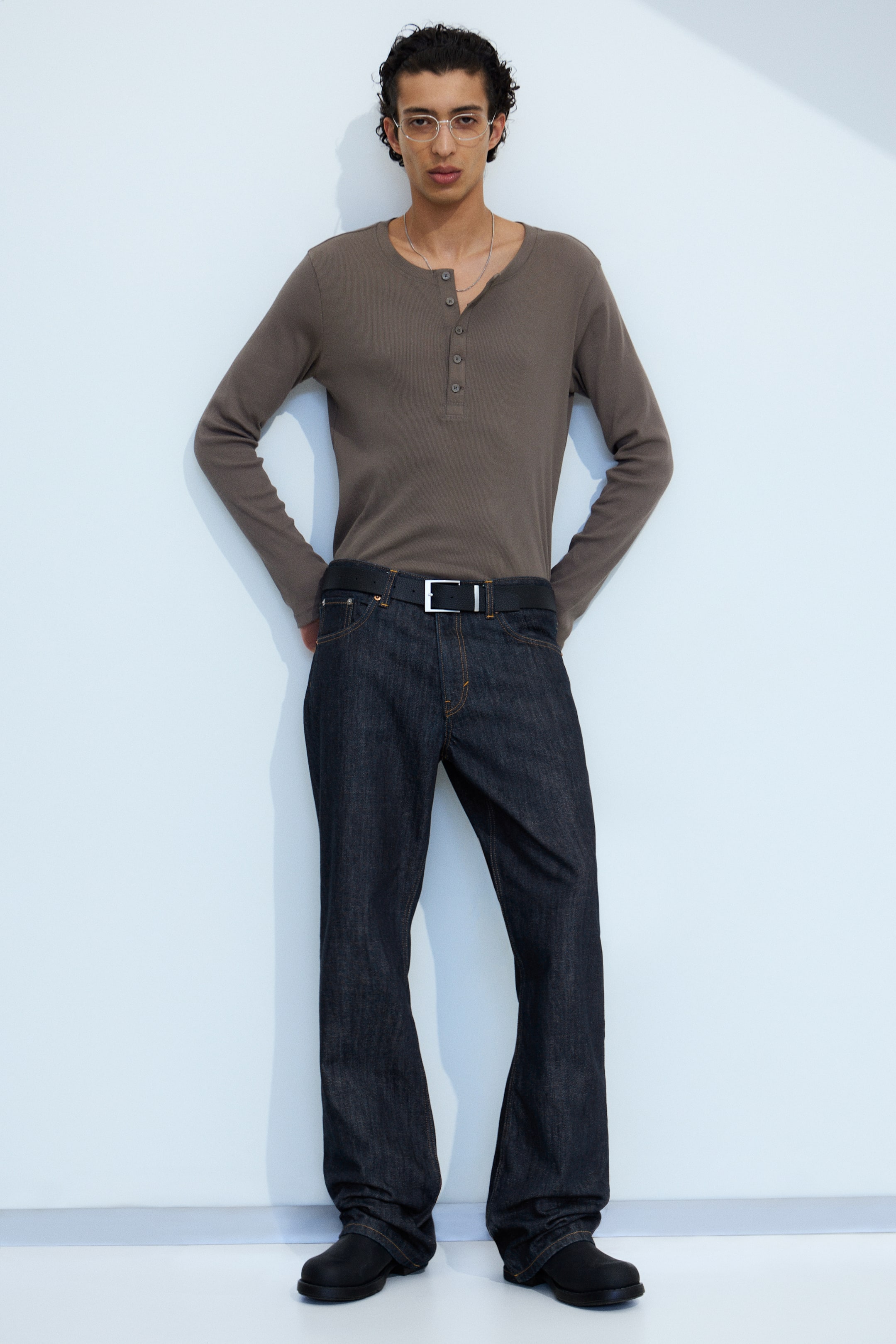 Ingrandisci l'immagine: Una maglia henley marrone a maniche lunghe, caratterizzata da un'allacciatura a quattro bottoni, è indossata infilata in un paio di jeans scuri. I jeans scuri presentano una vestibilità rilassata con un leggero svasamento all'orlo, fissati da una cintura nera con fibbia argentata, e sono abbinati a scarpe nere.