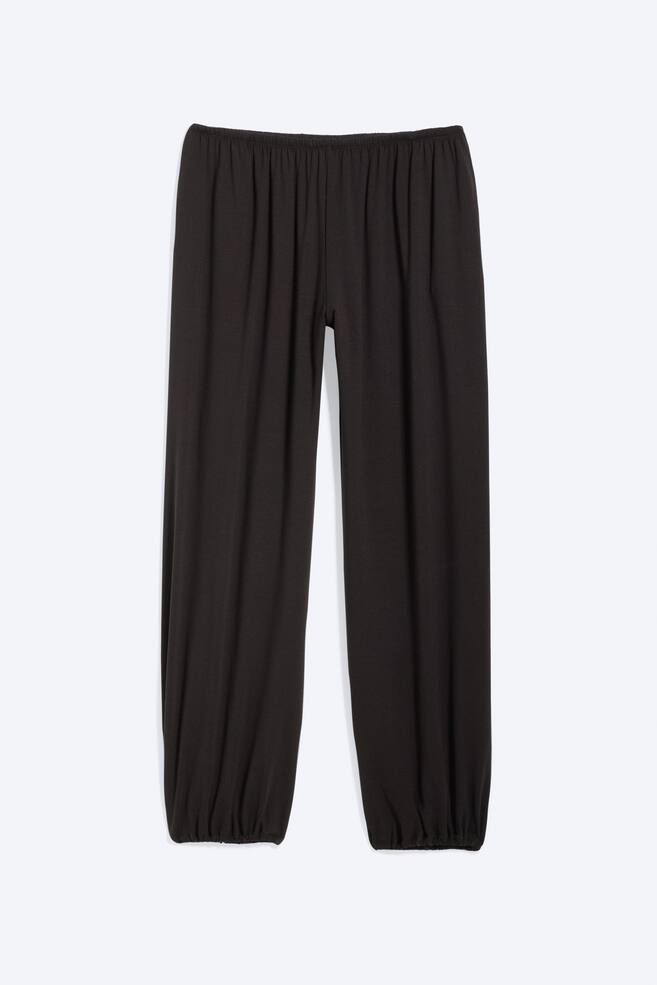 A pair of black trousers is displayed from the front, showcasing an elasticated, gathered waistband. The trousers are designed with loose, balloon-shaped legs that gather at the elasticated cuffs. - 1
