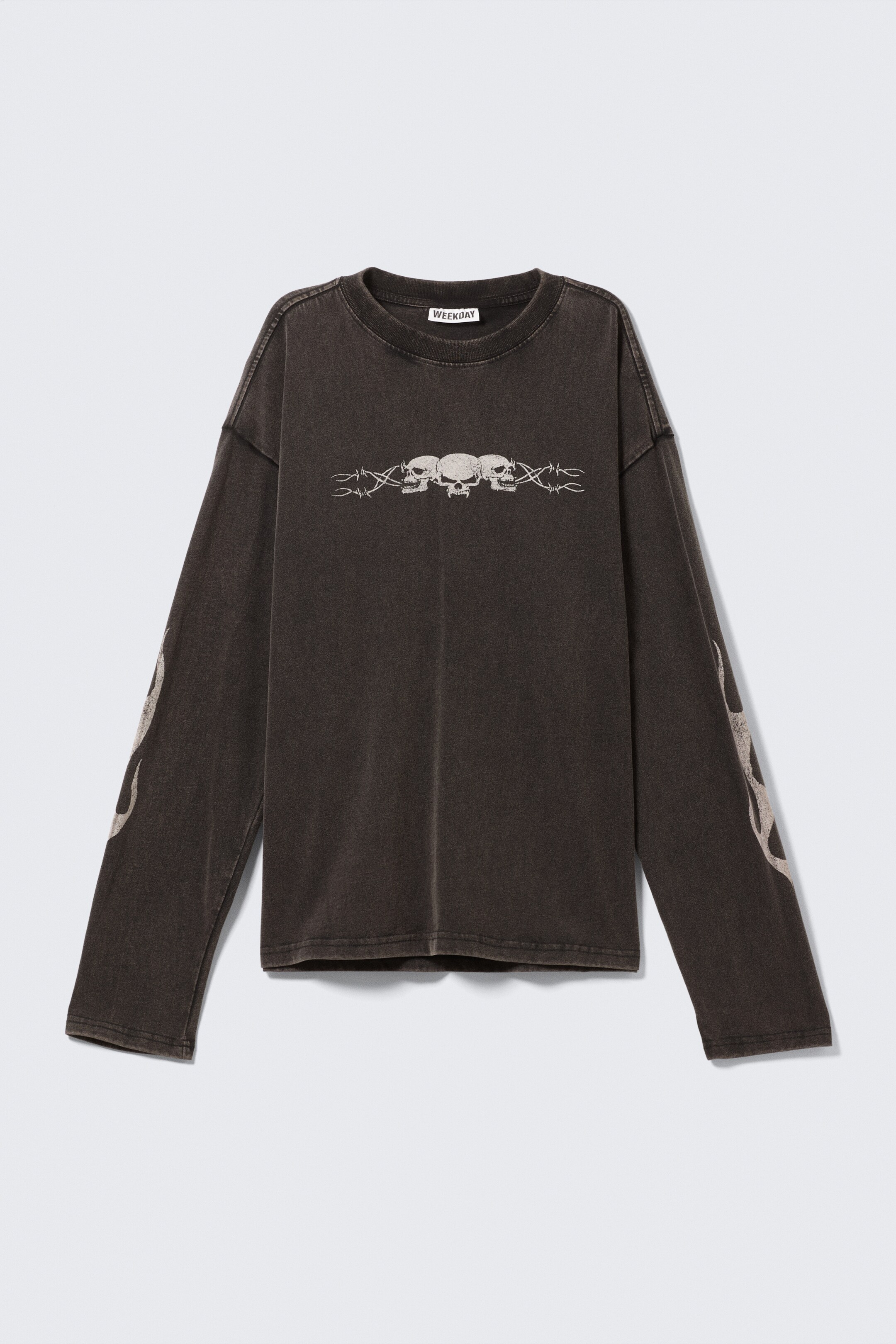 Ingrandisci l'immagine: Una T-shirt a maniche lunghe in un grigio scuro lavato presenta il suo davanti, caratterizzata da una vestibilità regolare e squadrata e un collo rotondo a coste. La maglietta è adornata con grafiche di colore chiaro: tre teschi bordati da disegni di filo spinato sul petto e motivi di fiamme che corrono verticalmente lungo il lato esterno di entrambe le maniche lunghe.