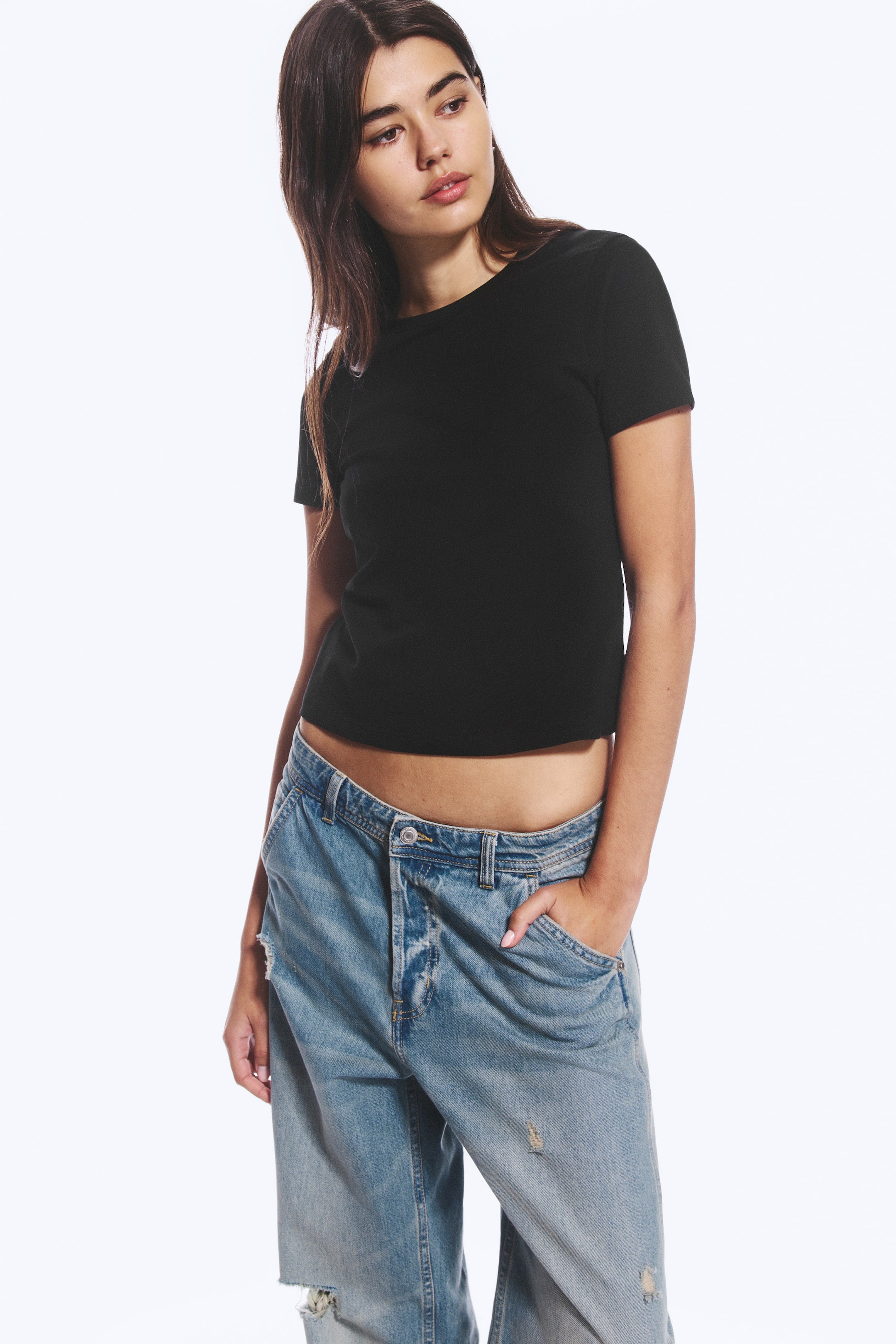 Grösseres Bild ansehen: Ein schwarzes, figurbetontes Mini-T-Shirt mit Rundhalsausschnitt und kurzen Ärmeln wird vom Model getragen. Das Model steht mit einer Hand in der Tasche einer distressed, hellblauen Jeans und betont dabei den verkürzten Saum des Shirts.
