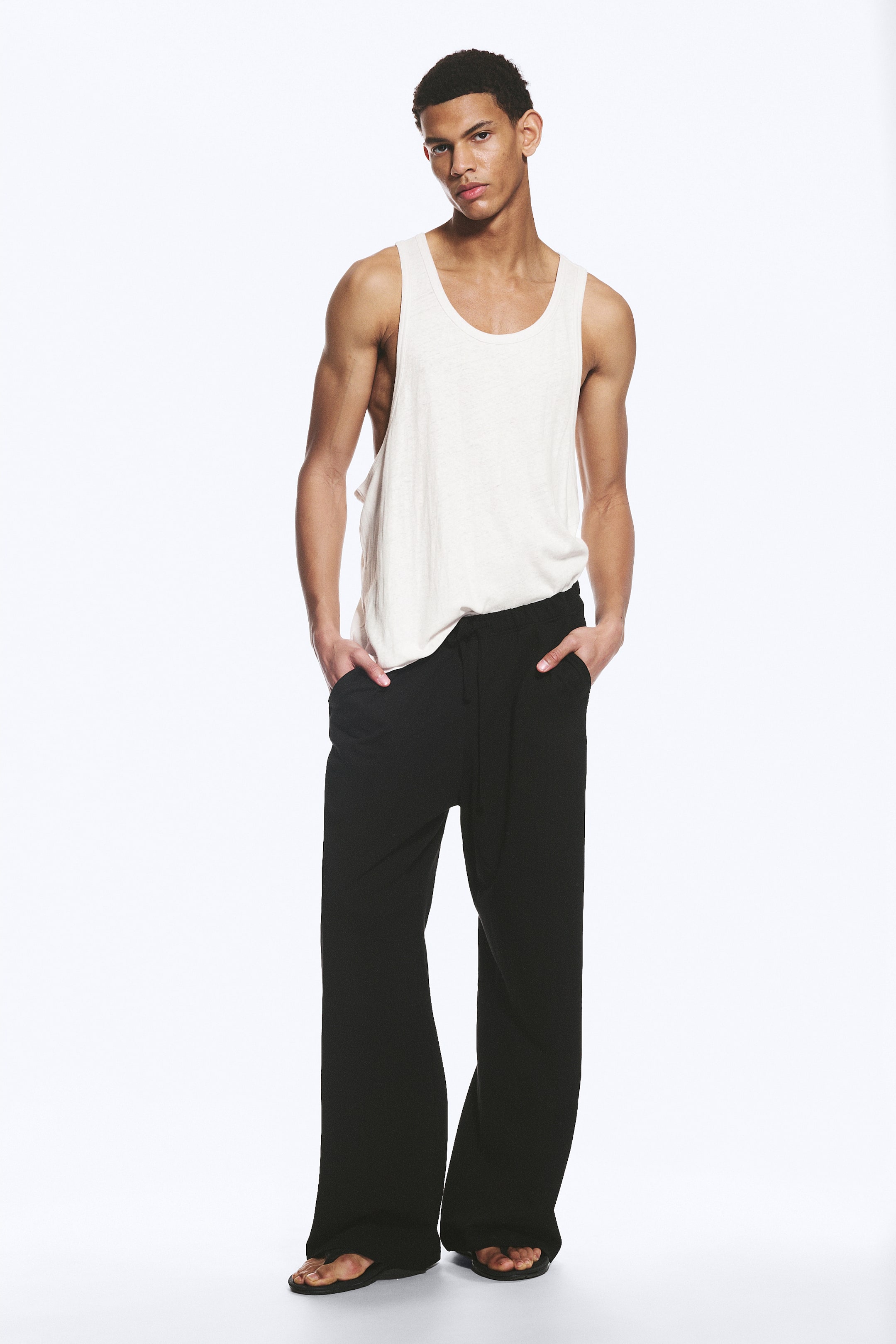 View larger image: An off-white vest top with a scoop neck and wide armholes is being worn, featuring a loose fit and a subtle texture. Black, wide-leg trousers are also being worn, featuring a drawstring waist, side pockets, and a relaxed drape that is pooling at the ankles.