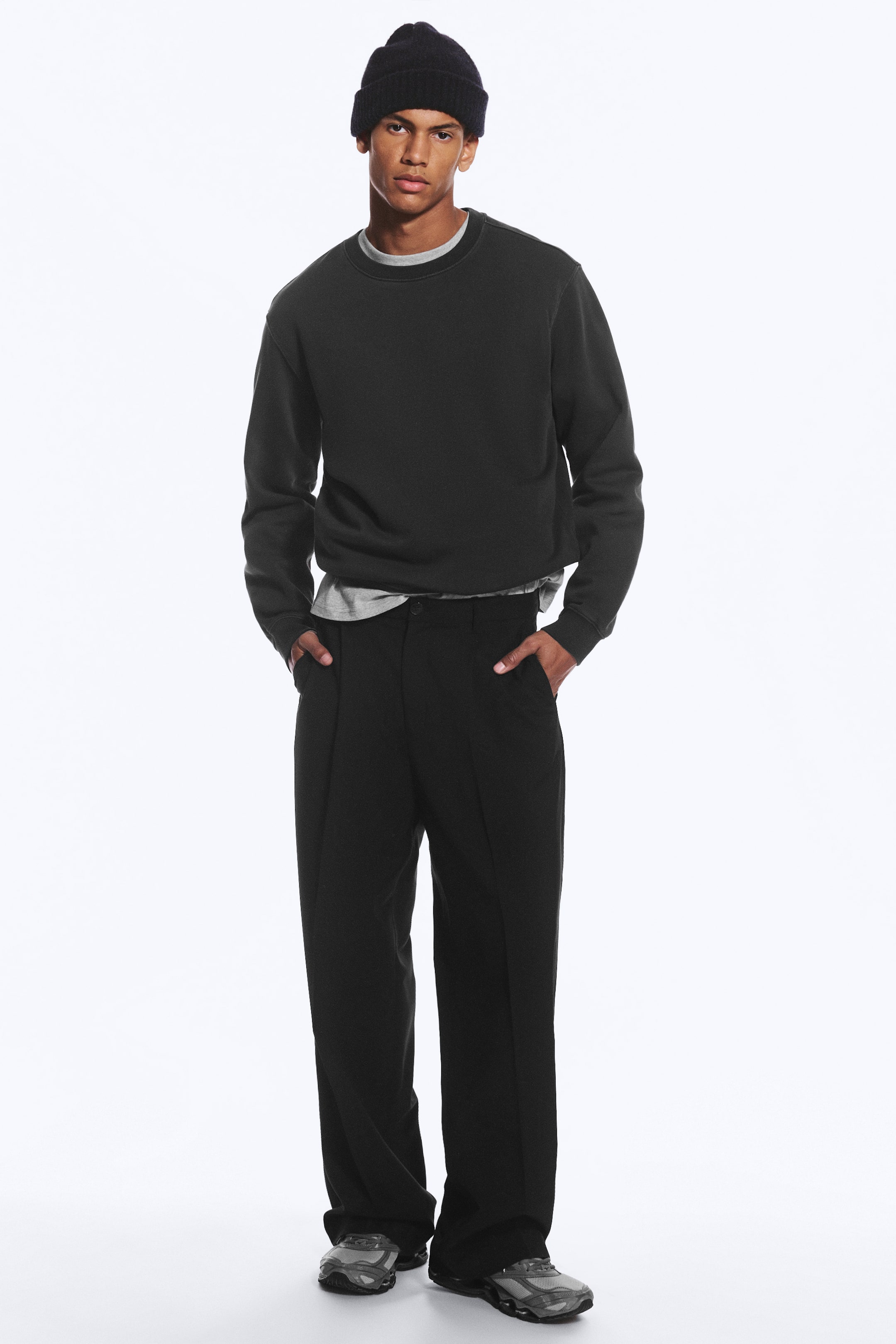 Vezi imaginea mai mare: Un model poartă o pereche de pantaloni croiți negri, oversize, cu pense în față și dungi presate, stând cu mâinile în buzunarele laterale. Pantalonii cu croială largă sunt asortați cu un hanorac închis la culoare cu decolteu rotund, un tricou gri pe dedesubt și o căciulă neagră.