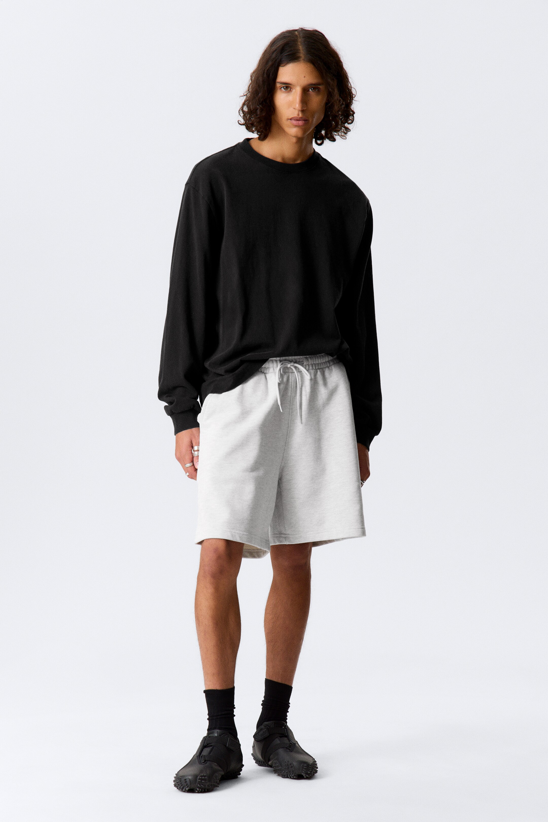 Grotere afbeelding bekijken: Een zwarte trui met lange mouwen en ronde hals heeft een ontspannen pasvorm en ronde halsstijl, gezien vanaf de voorkant. Hij wordt gedragen met lichtgrijze sweatshorts, die voorzien zijn van een elastische tailleband en een zichtbare witte trekkoord.