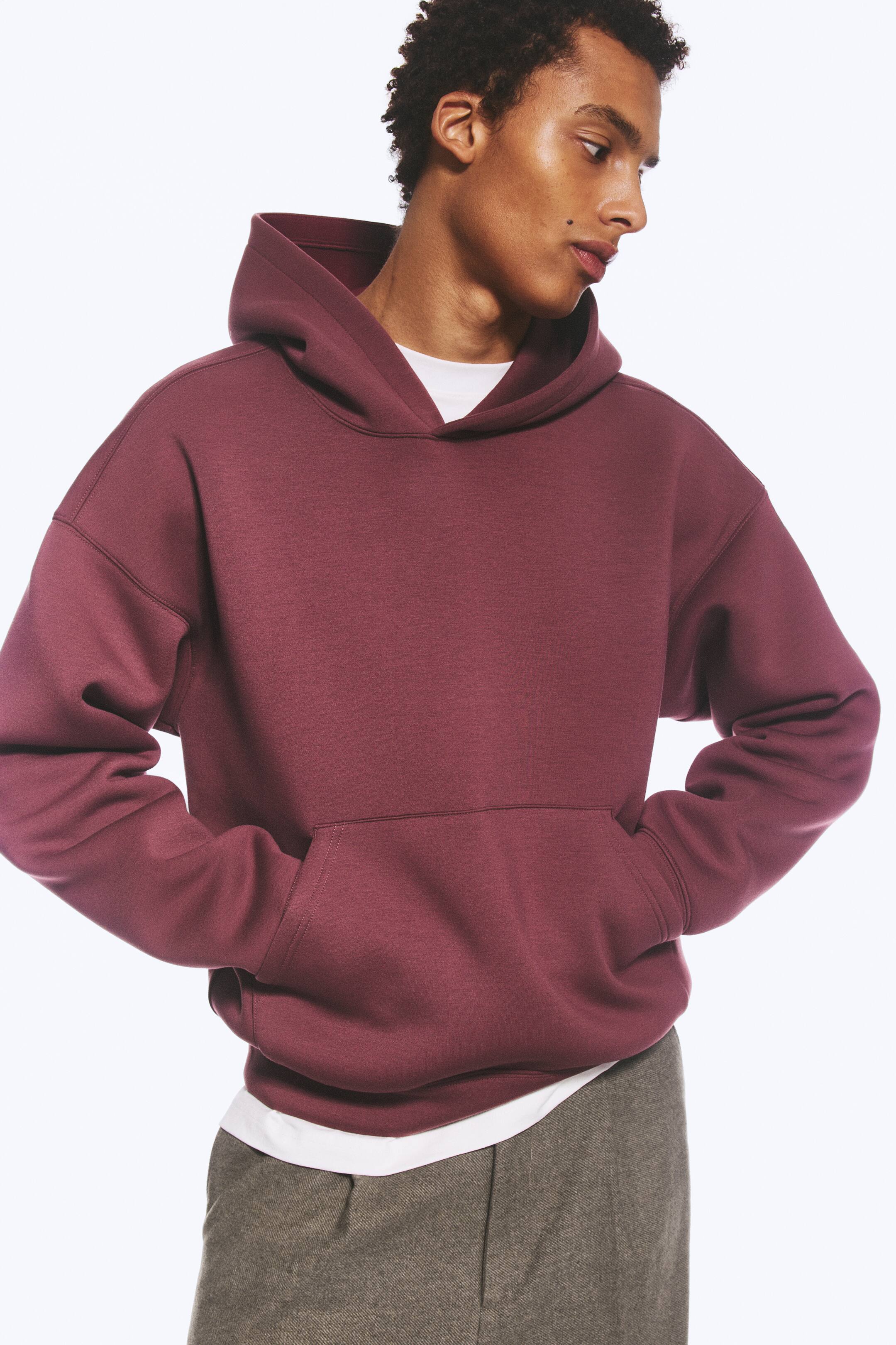 View larger image: A burgundy loose-fit hoodie in a structured scuba fabric is being worn by the model, who has both hands in the front kangaroo pocket. The hoodie features dropped shoulders and is layered over a white T-shirt.