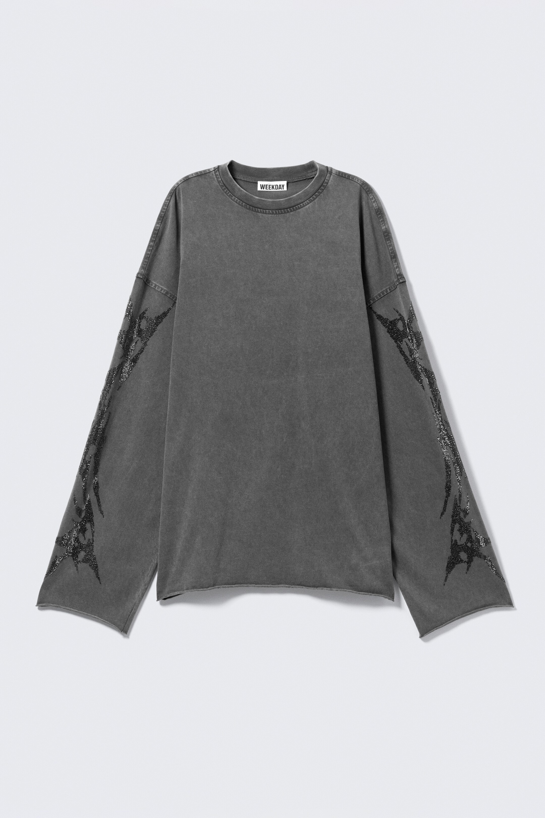 Grotere afbeelding bekijken: Een vooraanzicht van een oversized, donkergrijs T-shirt met lange mouwen, gemaakt van katoenen jersey, vertoont een gewassen uitstraling. Het heeft een ronde halslijn, een ruwe onderzoom en ruwe mouwboorden, met ingewikkelde rhinestone-patronen die de buitenzijden van beide mouwen sieren.