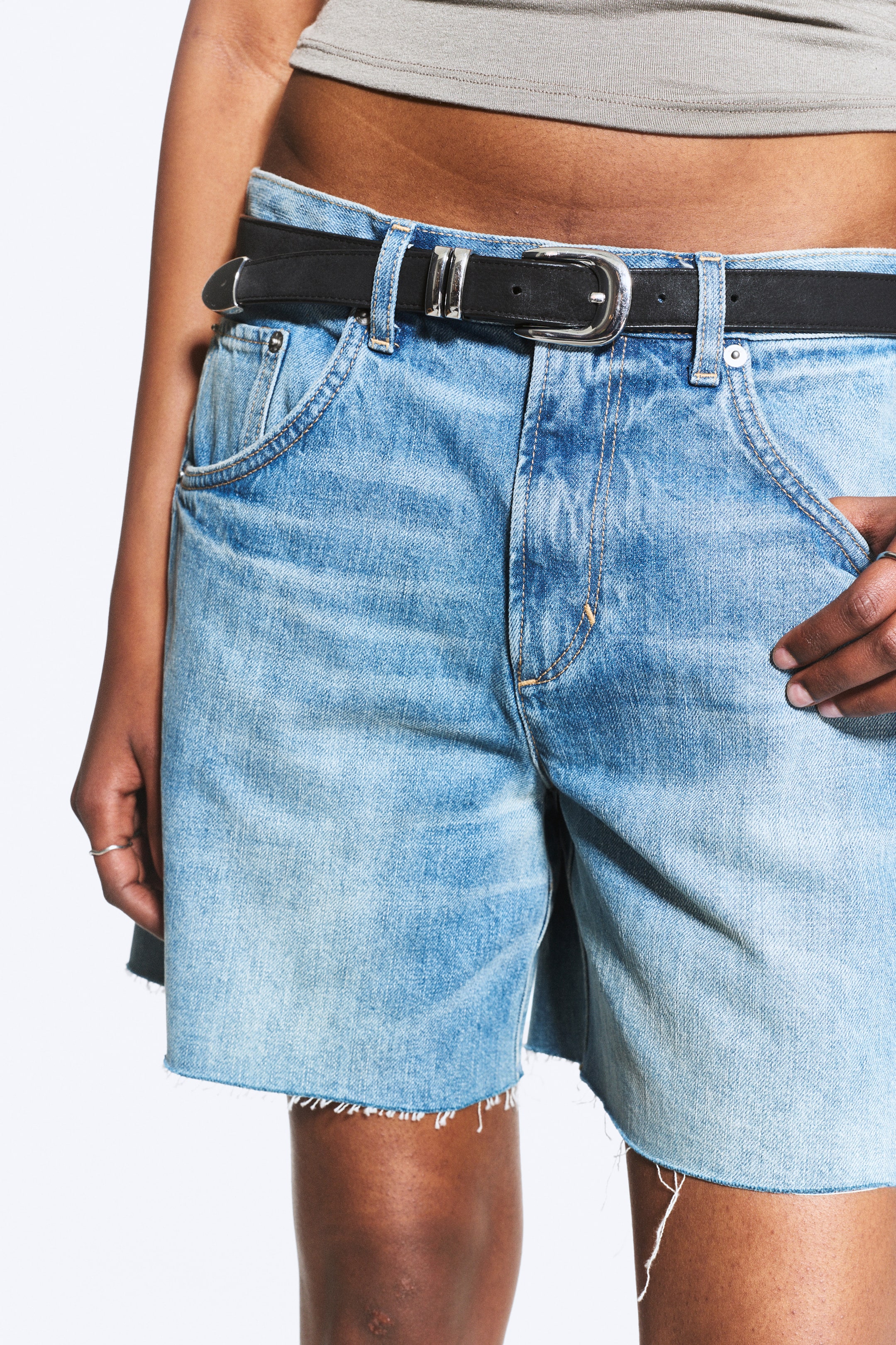 Größeres Bild anzeigen: Hell gewaschene Jeansshorts werden aus einem frontrechten Winkel präsentiert, mit einem verblassten Aussehen, rohen ausgefransten Säumen und sichtbaren Vordertaschen, einschließlich einer Münztasche. Ein schwarzer Gürtel mit einer silberfarbenen rechteckigen Schnalle und zwei Schlaufen ist durch die Gürtelschlaufen der Shorts gezogen.