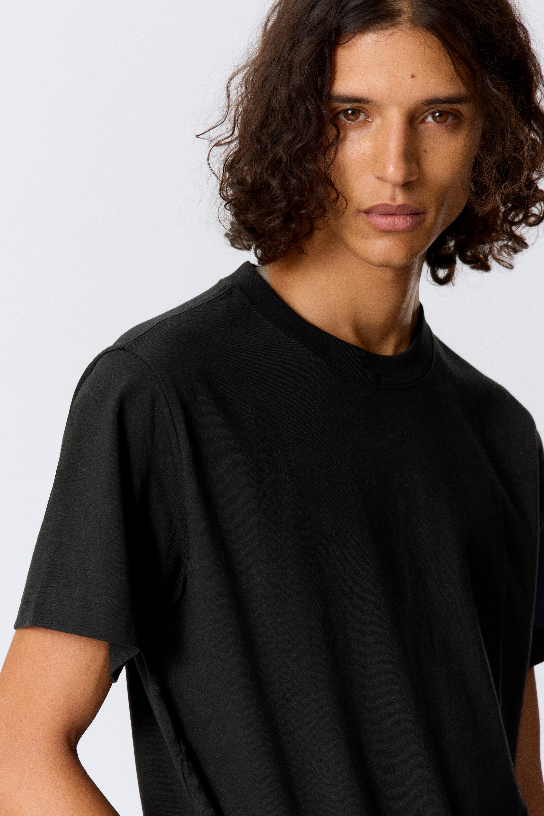 Ver imagem maior: Uma t-shirt preta de gola redonda está a ser usada, virada para a frente direita, com uma gola canelada, mangas curtas relaxadas e um tecido suave e drapeado.