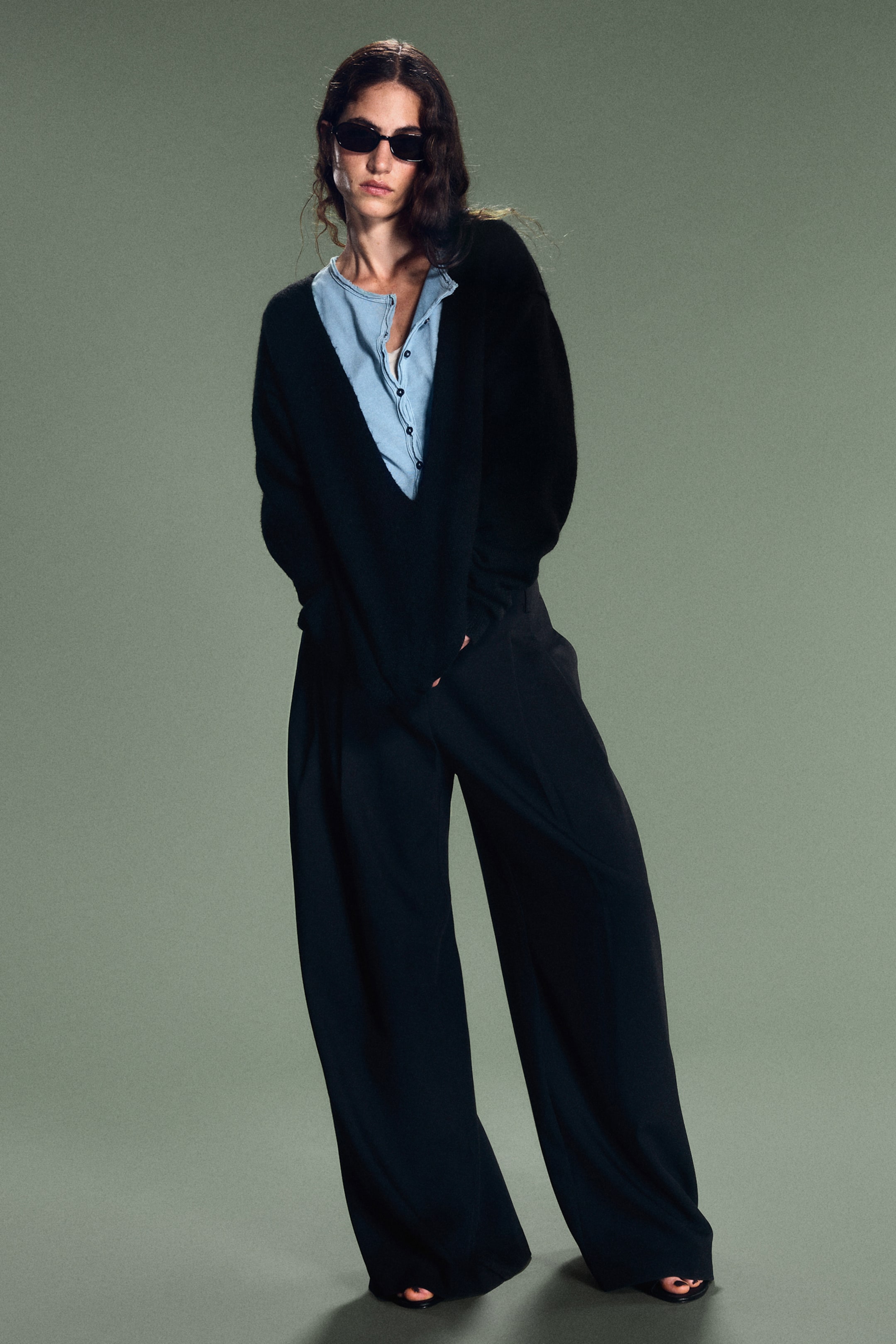 View larger image: Black wide-leg pleated suit trousers are being worn by the model, showing a relaxed fit with front-pleat detailing. The trousers are styled with an oversized black V-neck jumper layered over a light blue button-up shirt.