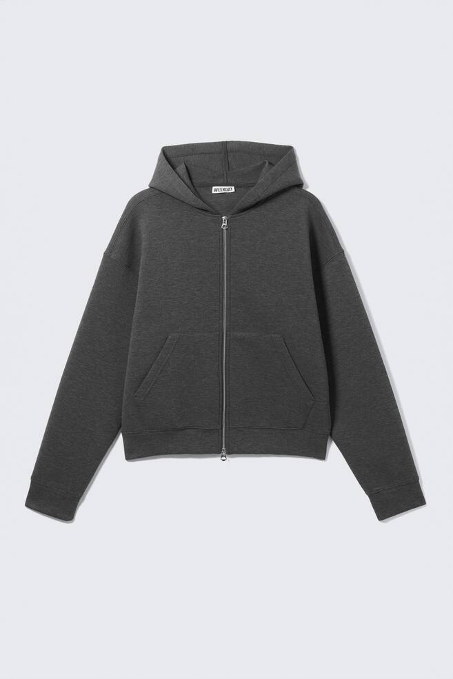 Ein dunkelgrauer Kapuzenpullover mit Reißverschluss ist von vorne zu sehen, mit einer dicken, glatten Textur, leicht überschnittenen Schultern, gerippten Bündchen und einem gerippten Saum. Ein silberfarbener Zwei-Wege-Reißverschluss verläuft mittig, flankiert von zwei eckigen Vordertaschen, mit einem 'WEEKDAY'-Label, das im inneren Nackenbereich sichtbar ist. - 1