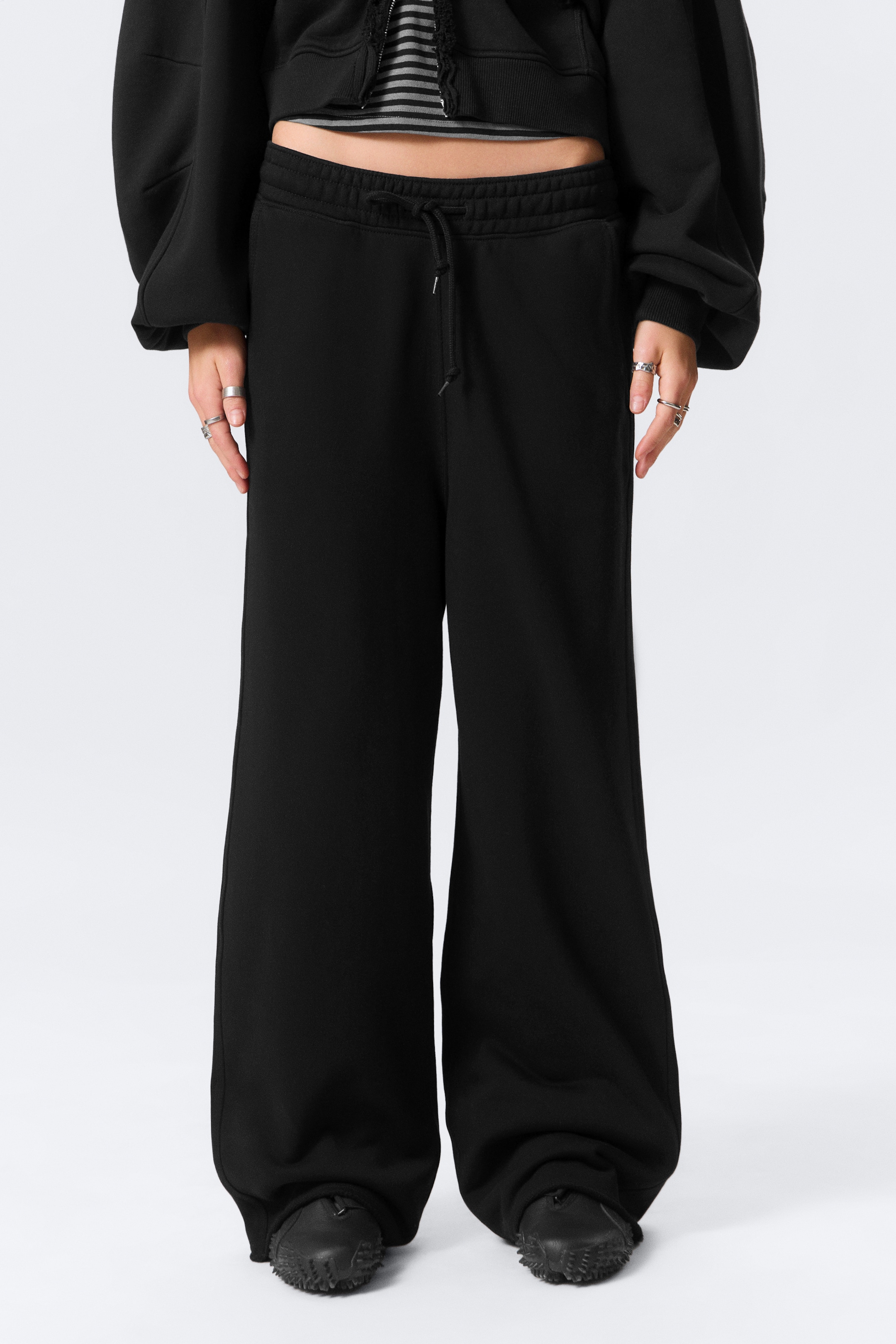 Visualizza immagine più grande: I pantaloni della tuta neri a gamba larga sono presentati da una vista frontale, con una cintura elastica e un cordoncino annodato. I pantaloni mostrano una vestibilità rilassata, con le gambe larghe che si accumulano intorno ai piedi di chi li indossa.