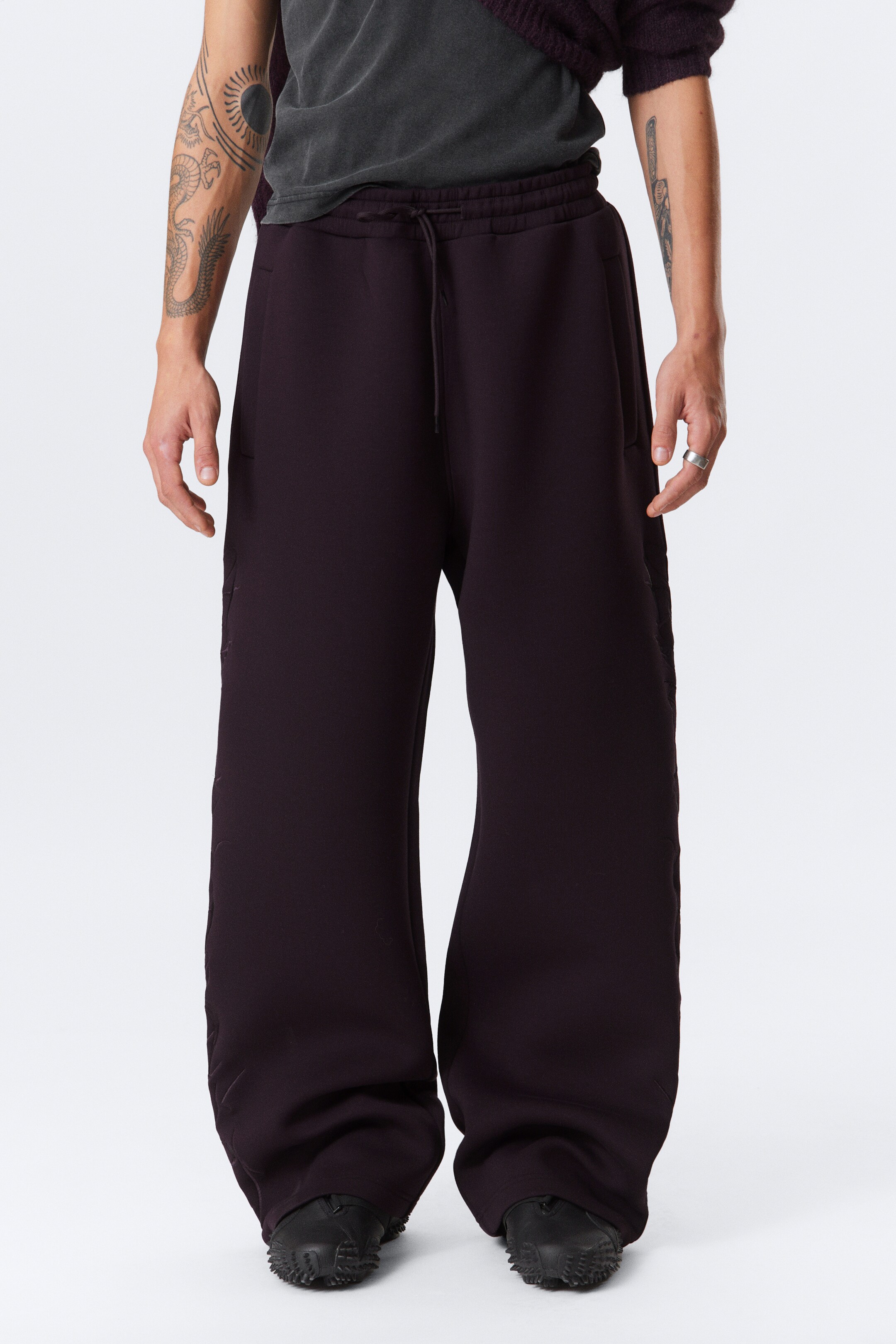Agrandir l'image: Le pantalon de jogging large couleur prune foncé est porté de face, avec une ceinture élastiquée avec cordon de serrage et des poches latérales discrètes. Le tissu ample s'accumule autour des pieds de la personne qui le porte.