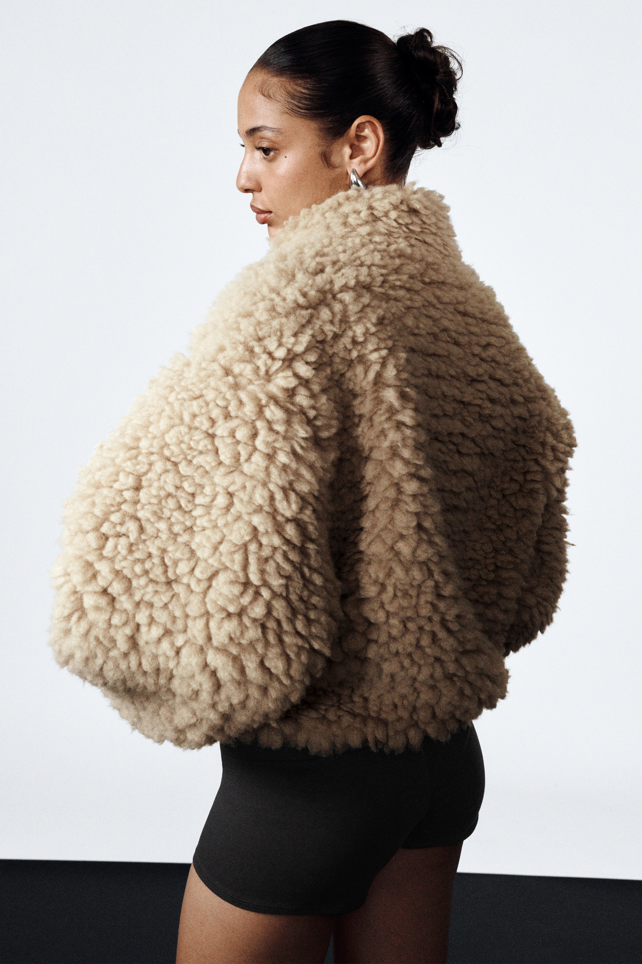 View larger image: A light beige curly pile jacket is being presented from a side and back angle, showcasing its voluminous, cropped silhouette over dark shorts. The jacket features a high, standing collar and wide sleeves.