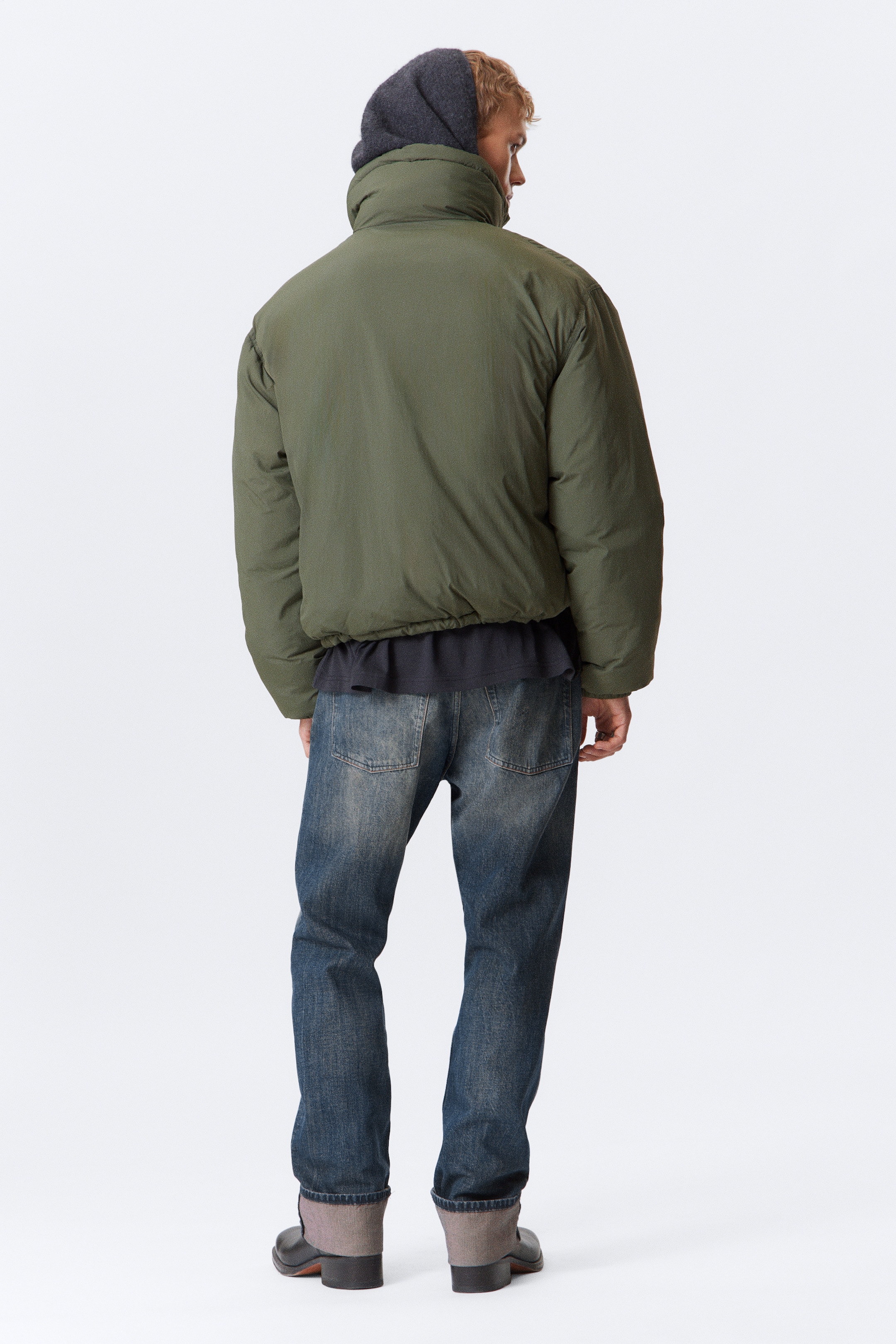 Visualizza immagine più grande: Una giacca imbottita trapuntata verde oliva con un collo a imbuto alto, polsini elasticizzati e una silhouette corta e ampia è indossata dalla modella. Il suo orlo arricciato rivela una maglietta scura sotto, e un indumento con cappuccio scuro è visibile sotto il collo.