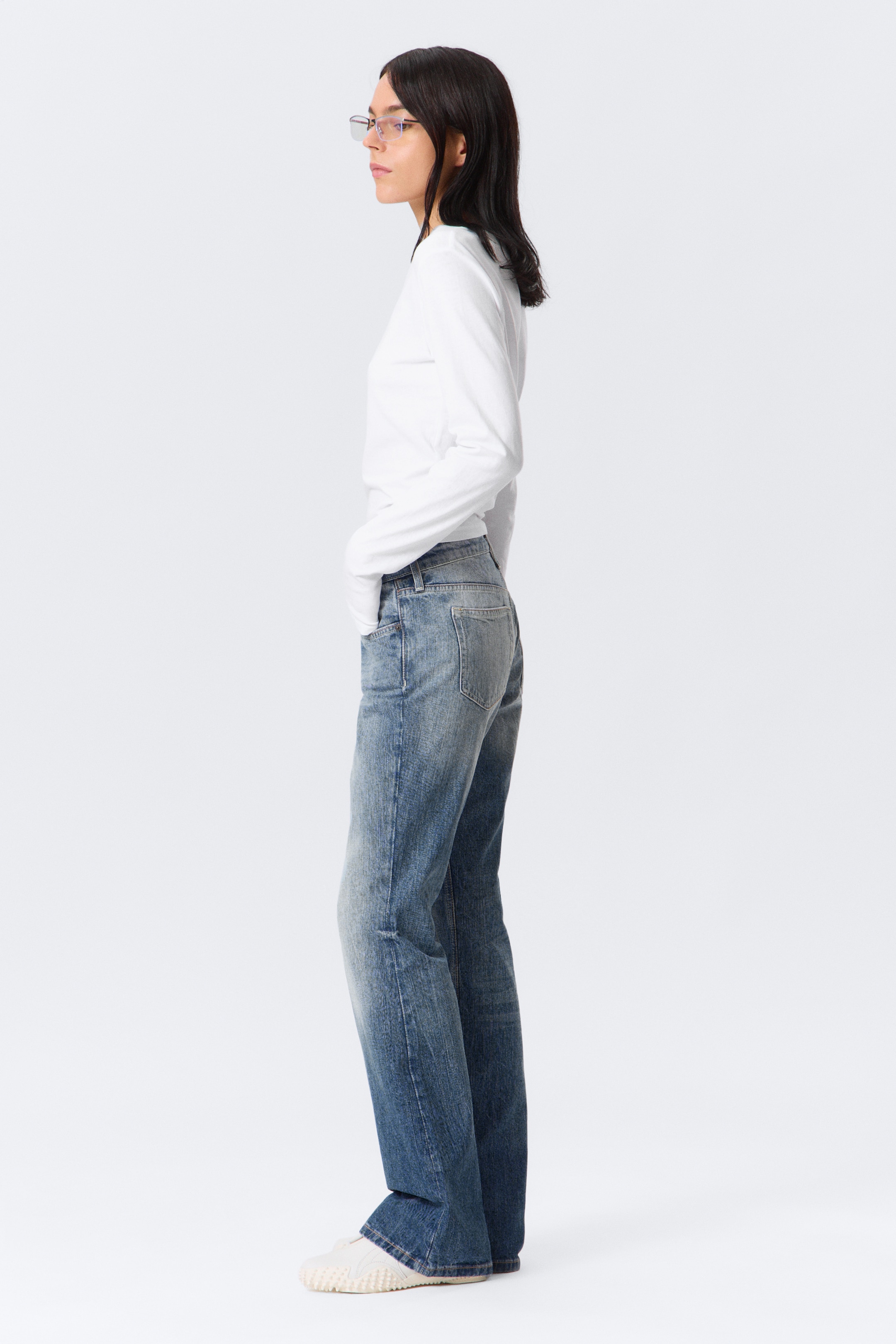 Grotere afbeelding bekijken: Een wit lang mouwloos top en blauwe jeans worden gepresenteerd op een persoon die in profiel staat, naar links gericht. Het slim-fit topje is effen wit, terwijl de blauwe jeans een medium wassing hebben met zichtbare vervaging, whiskering en achterzakken, gesneden in een rechte pijp stijl die lichtgekleurde lage sneakers bedekt.