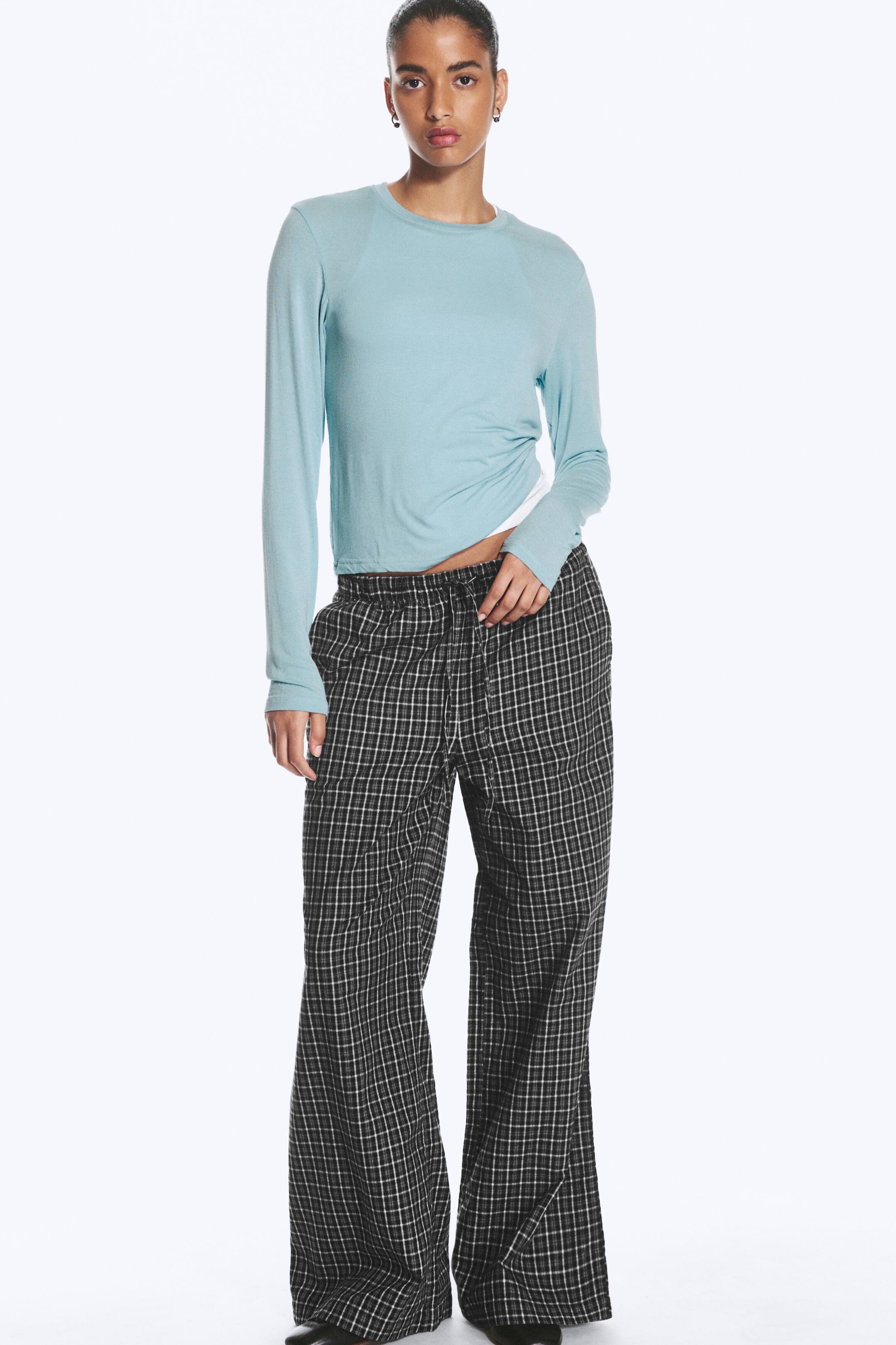 Größeres Bild ansehen: Das Model trägt eine weit geschnittene Flanell-Pyjamahose mit schwarz-weißem Karomuster. Die Hose verfügt über einen mittelhohen, elastischen Bund mit Kordelzug und wird mit einem hellblauen Langarmshirt kombiniert.