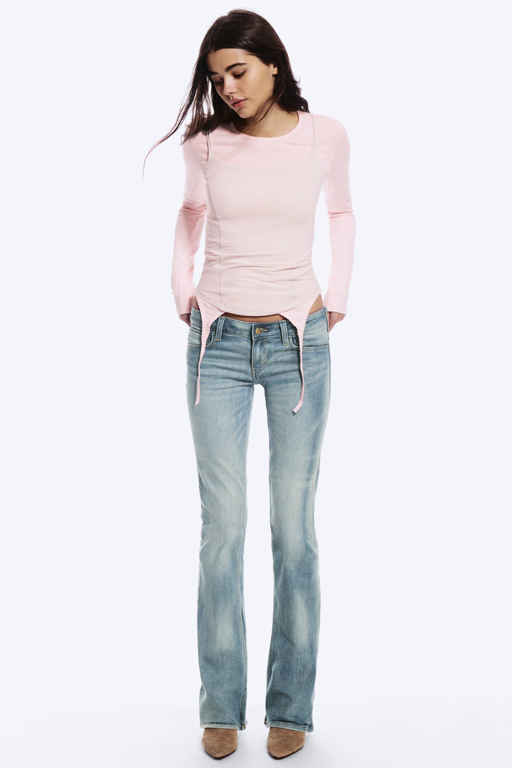 Visa större bild: Modellen bär ljus tvätt, lågmidjade bootcut-jeans och står med händerna vilande nära bakfickorna. De smala denimbyxorna har blekning längs låren och är stylade med en ljusrosa långärmad topp och bruna skor med spetsig tå.
