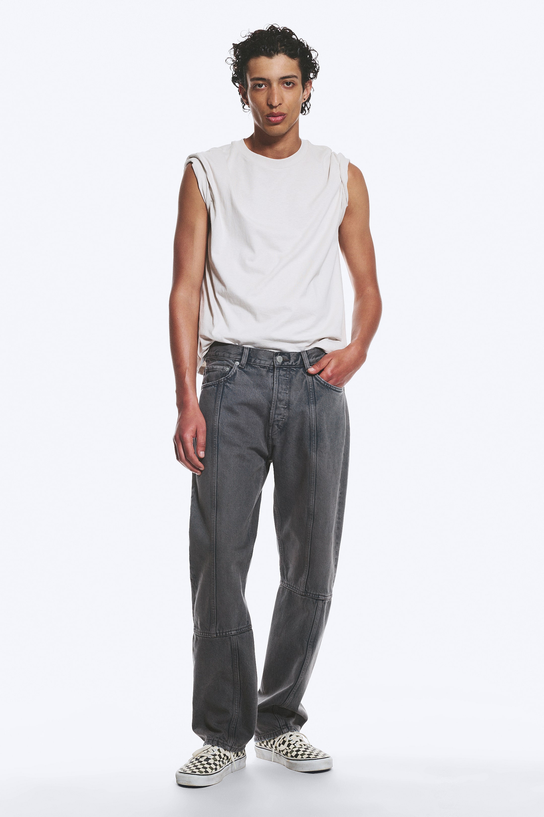 Grösseres Bild ansehen: Dunkelgraue Jeans mit entspannter Passform werden vom Model getragen, mit geradem Bein und sichtbaren Nahtdetails entlang der Vorderseite. Die Jeans verfügen über einen Knopfverschluss und ein 5-Taschen-Design, wobei die Hand des Models in einer Vordertasche ruht.