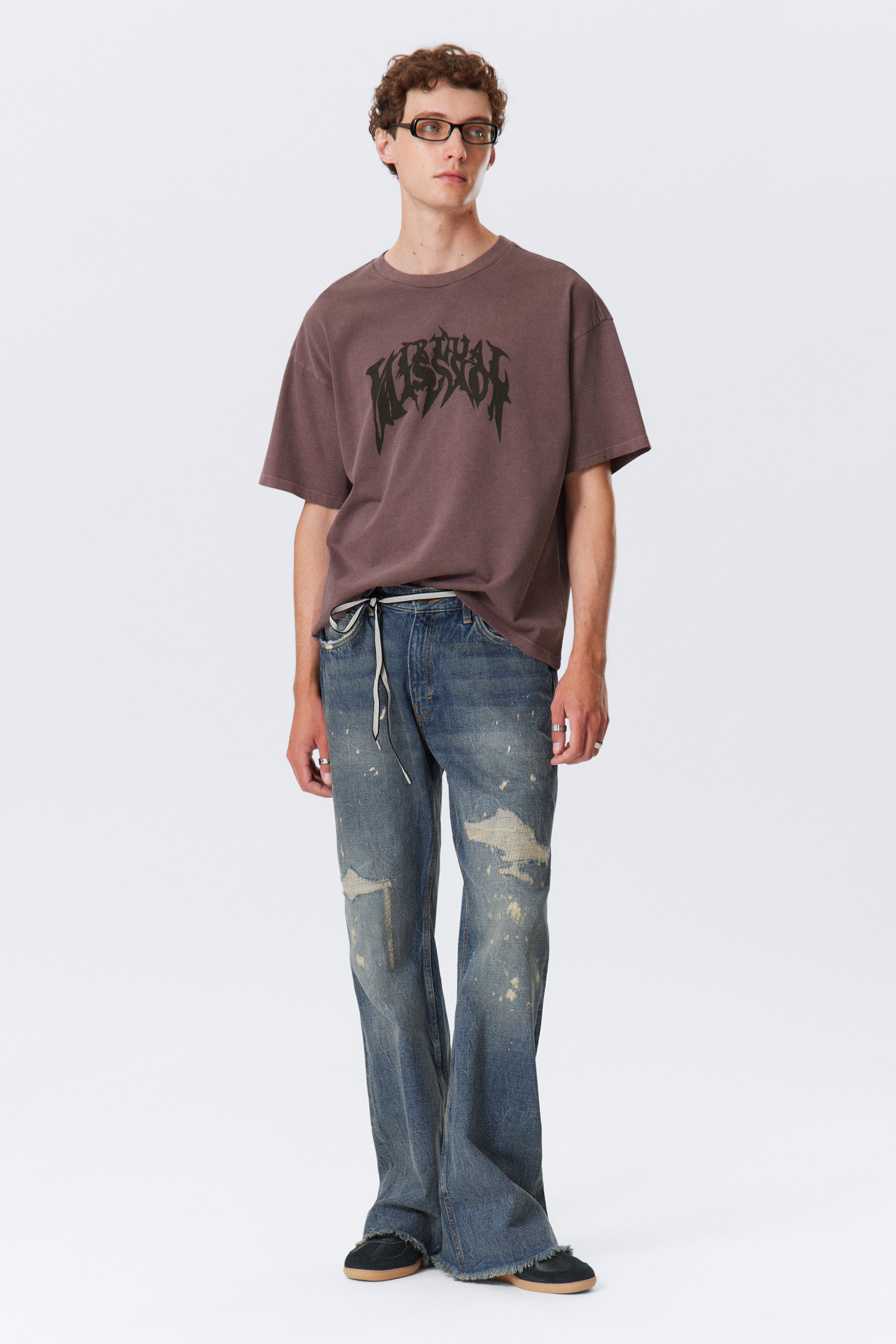 Größeres Bild anzeigen: Ein übergroßes, gedämpft braunes T-Shirt mit einem dunklen, abgenutzten Grafikdruck und einem Rundhalsausschnitt wird getragen, teilweise in eine locker sitzende, weit geschnittene blaue Jeans gesteckt. Die Jeans sind sichtbar abgenutzt mit Rissen, Farbspritzern und einem rohen, ausgefransten Saum und werden mit einem weißen Kordelzug gesichert, wobei schwarze Low-Top-Sneaker mit Gummisohlen das Ensemble vervollständigen.