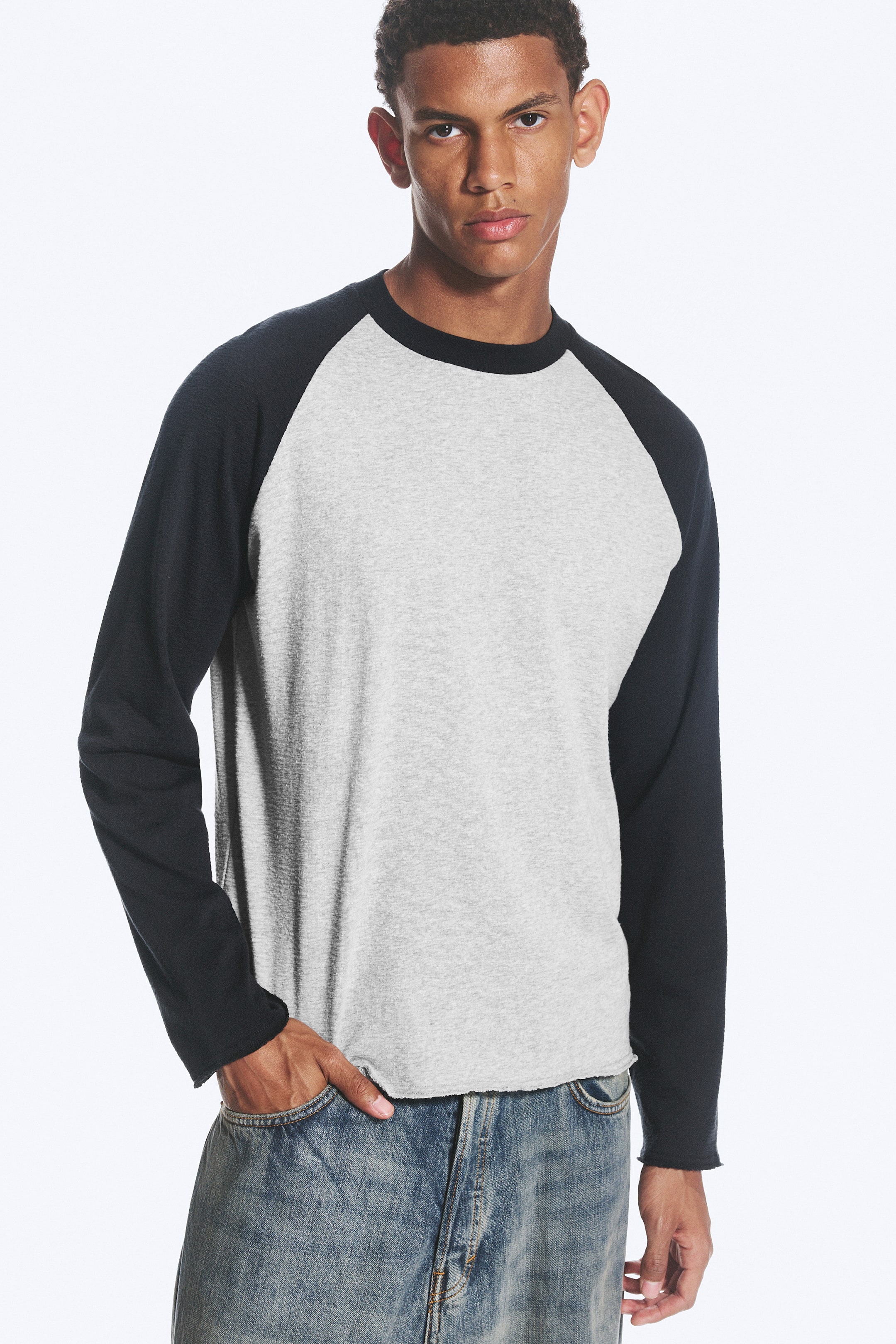 View larger image: A raglan long-sleeve shirt, featuring a grey body, black raglan sleeves, a black crew neck, and a raw bottom hem, is being worn by the model. The model is standing with one hand in a jeans pocket, pulling the shirt's hem.