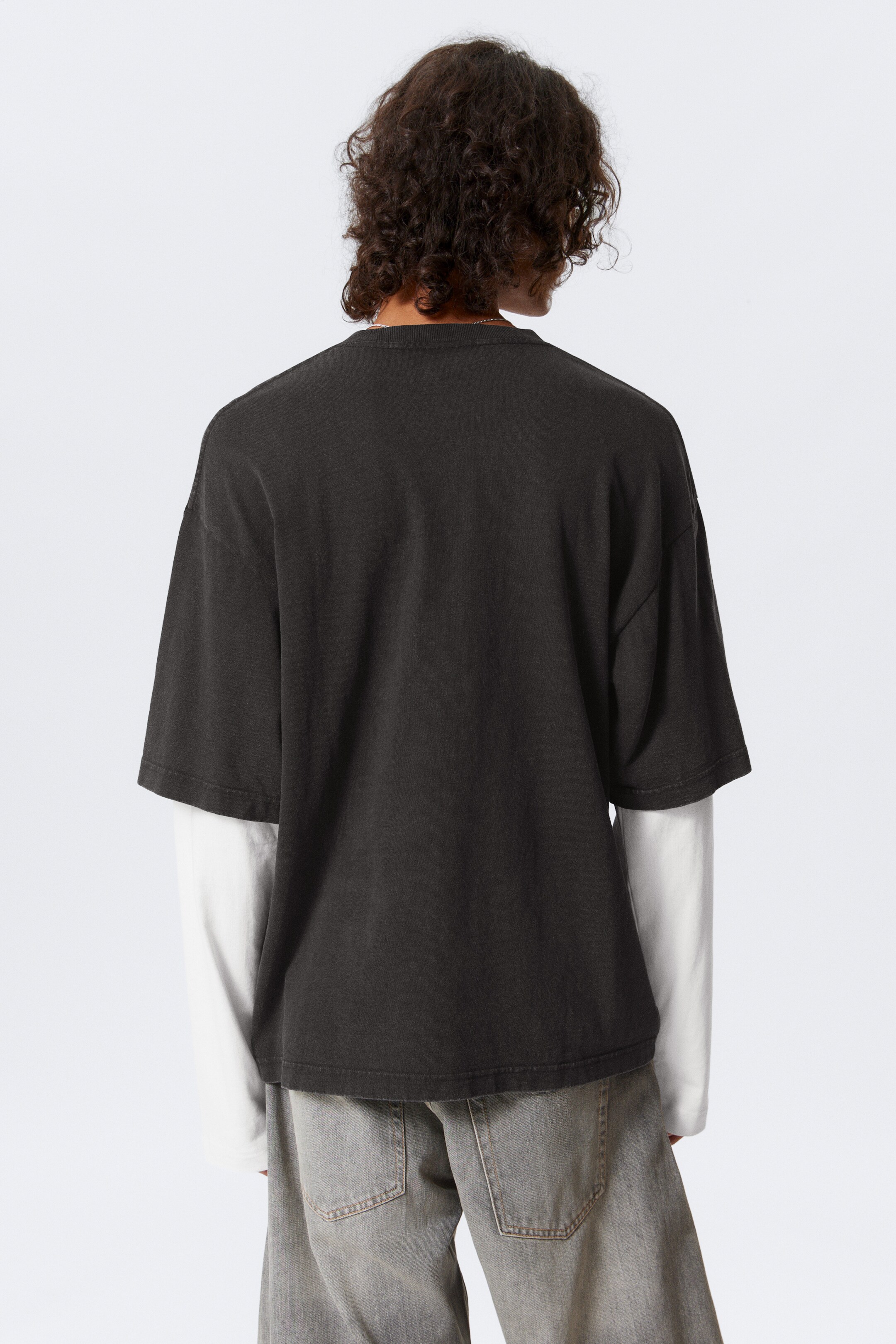View larger image: A dark, oversized short-sleeve t-shirt is being shown from the back, layered over a white long-sleeve shirt, with grey relaxed-fit pants also visible.