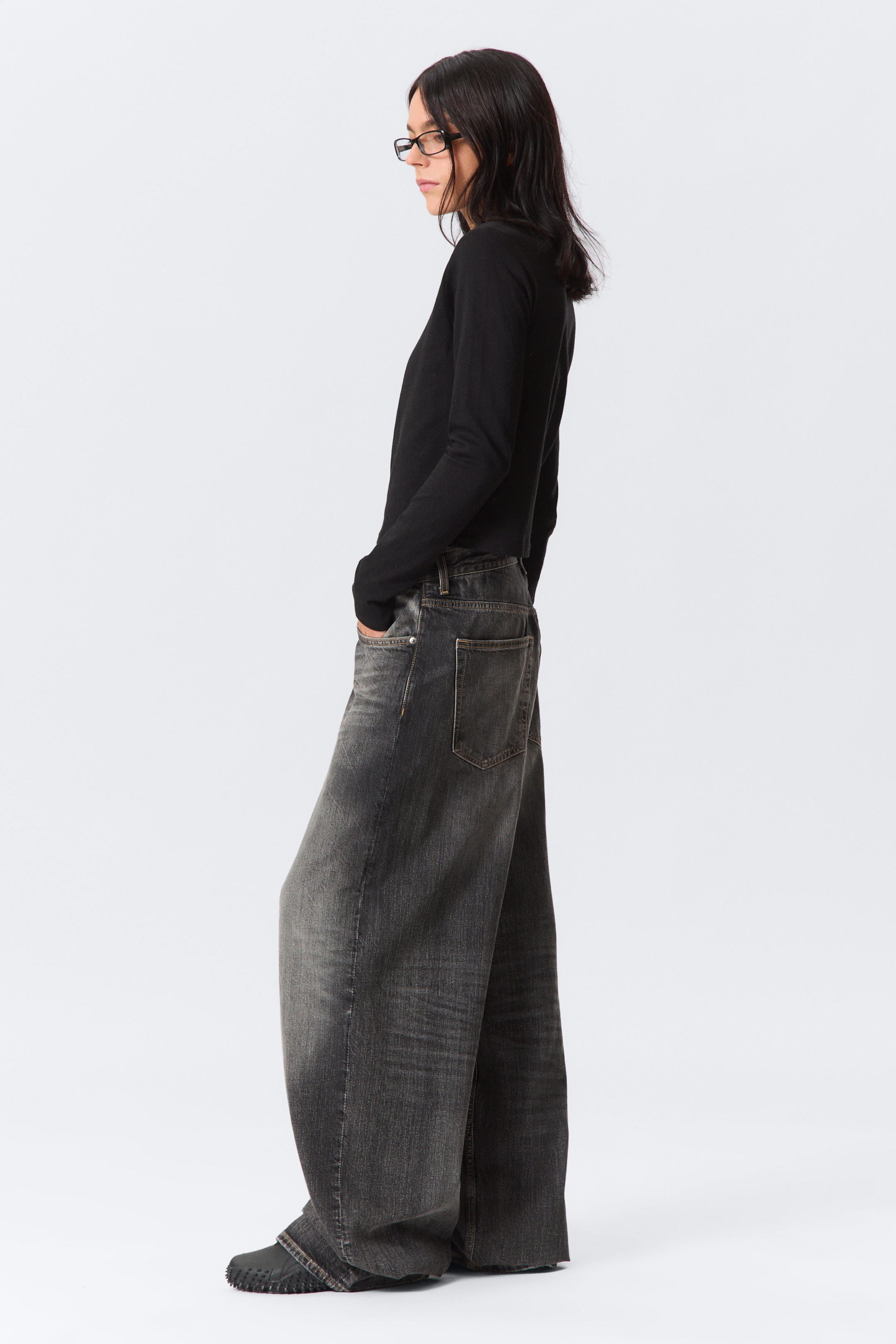 Ingrandisci l'immagine: Un top nero a maniche lunghe è indossato con jeans neri lavati a gamba larga, presentati di profilo. I jeans presentano una vestibilità rilassata e una texture leggermente sbiadita, con una tasca posteriore visibile.