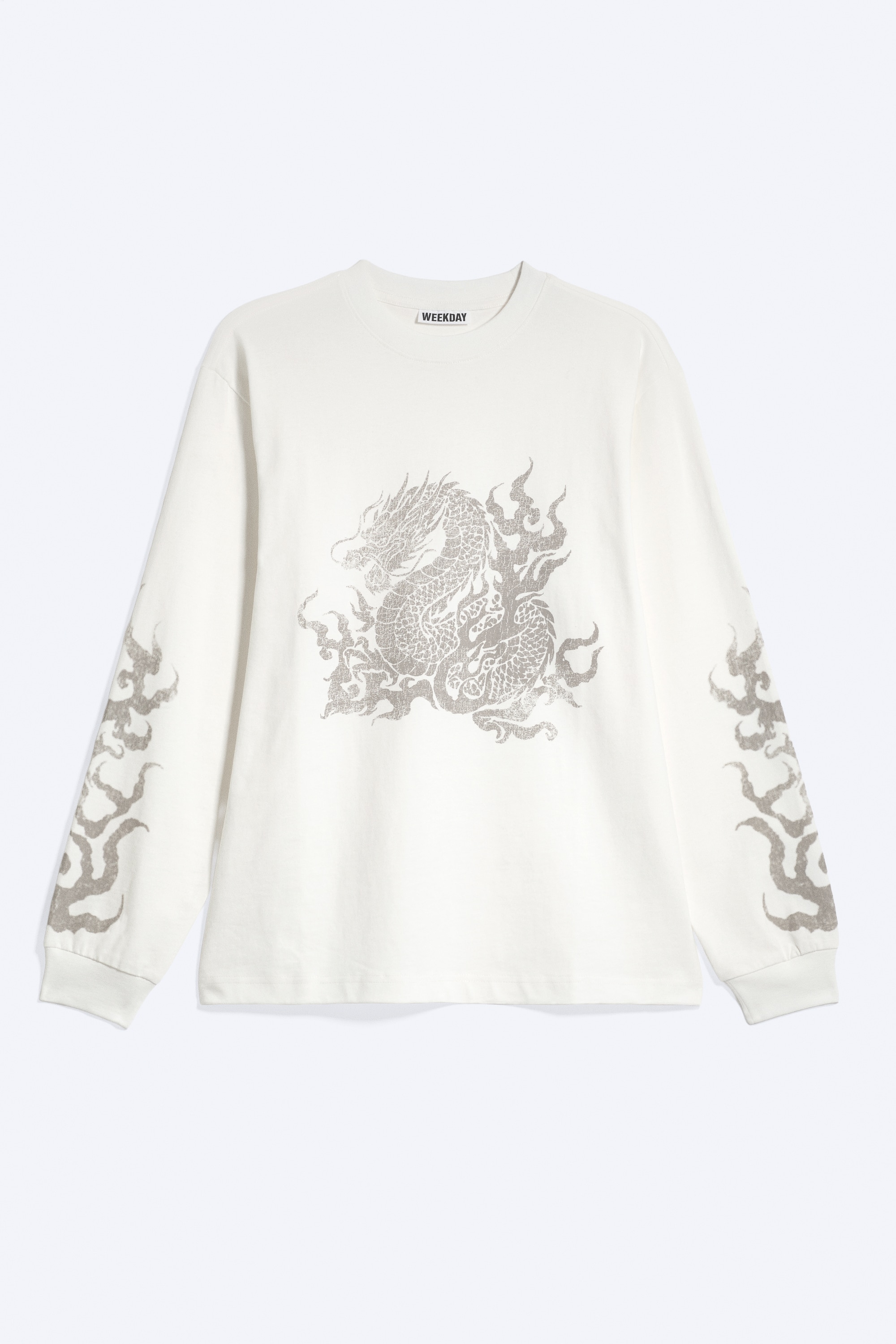 Un T-shirt blanc à manches longues orné d’un motif de dragon gris sur la poitrine et d’imprimés façon flammes sur les manches est porté par le modèle. Ce T-shirt à col rond et coupe décontractée est associé à une chemise à carreaux nouée autour de la taille.